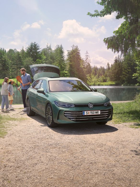 Der neue VW Passat Elegance in Maripositgrün Metallic parkt an einem Badesee mit geöffnetem Kofferraum. Ein Mann verteilt Freitzeitspielzeug an zwei Kinder.