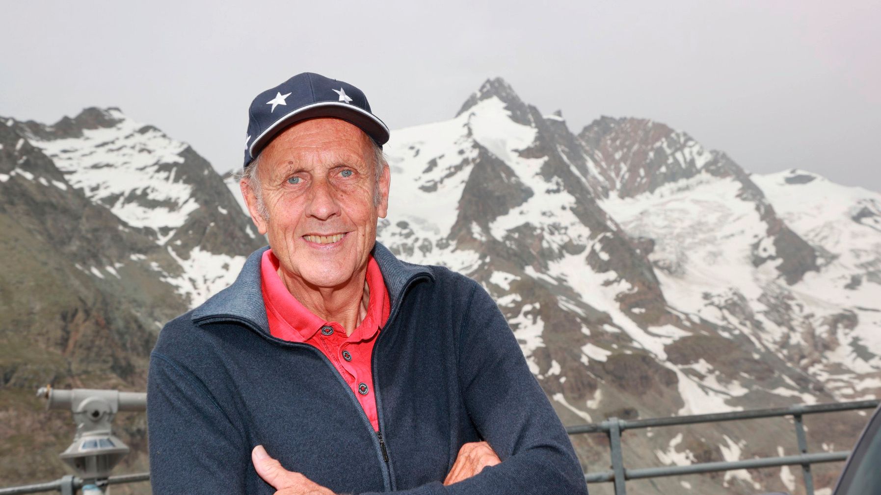 Rennsportlegende Hans-Joachim „Strietzel“ Stuck steht mit verschränkten Armen vor einer Berglandschaft mit schneebedeckten Gipfeln. Im Hintergrund sind Metallgeländer und ein Auto zu sehen. Der Himmel ist bewölkt.
