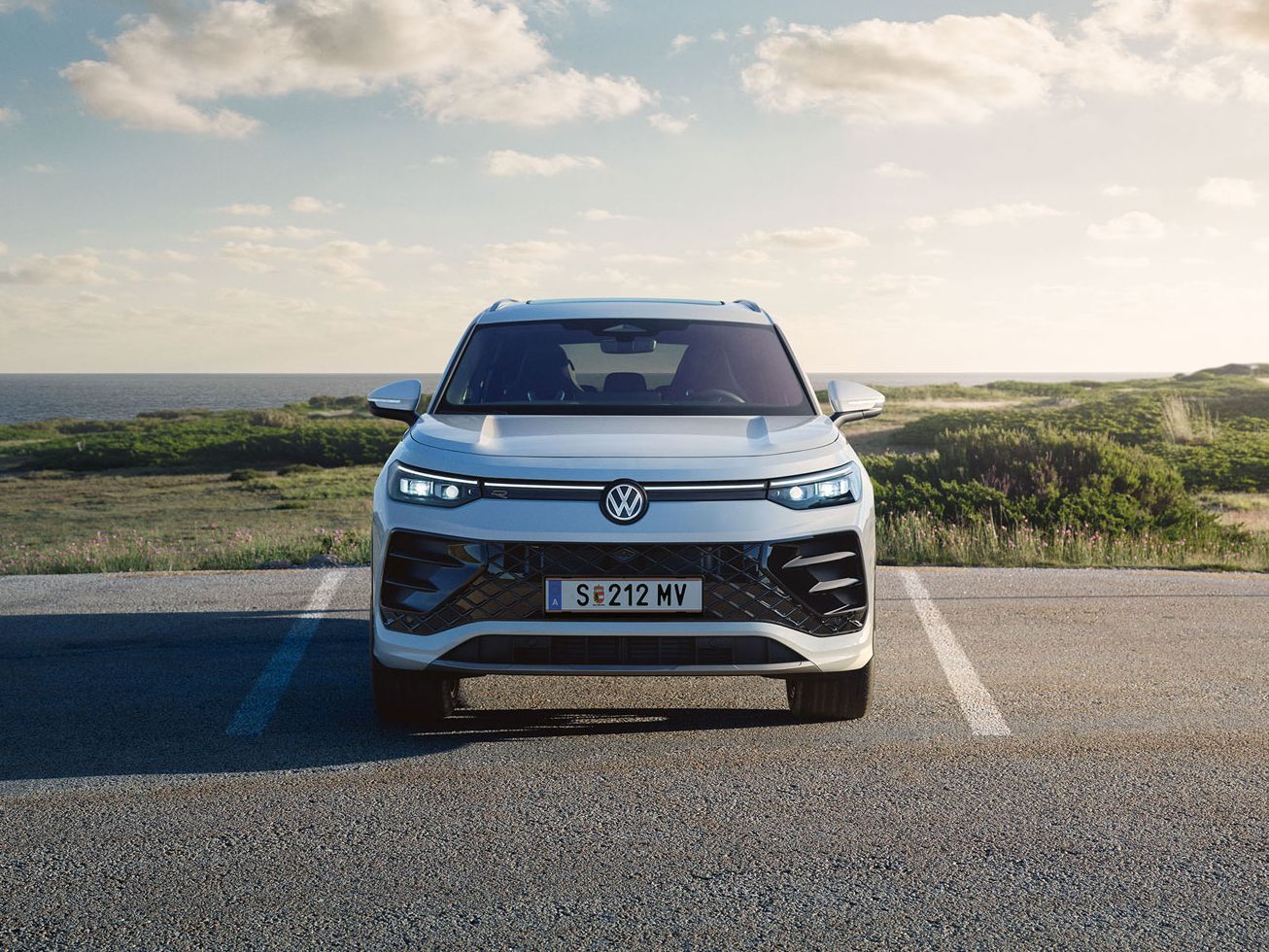 Frontansicht eines weißen VW Tayron auf einem Parkplatz in Dünen, die  Front-Lichtsignatur ist angeschaltet.