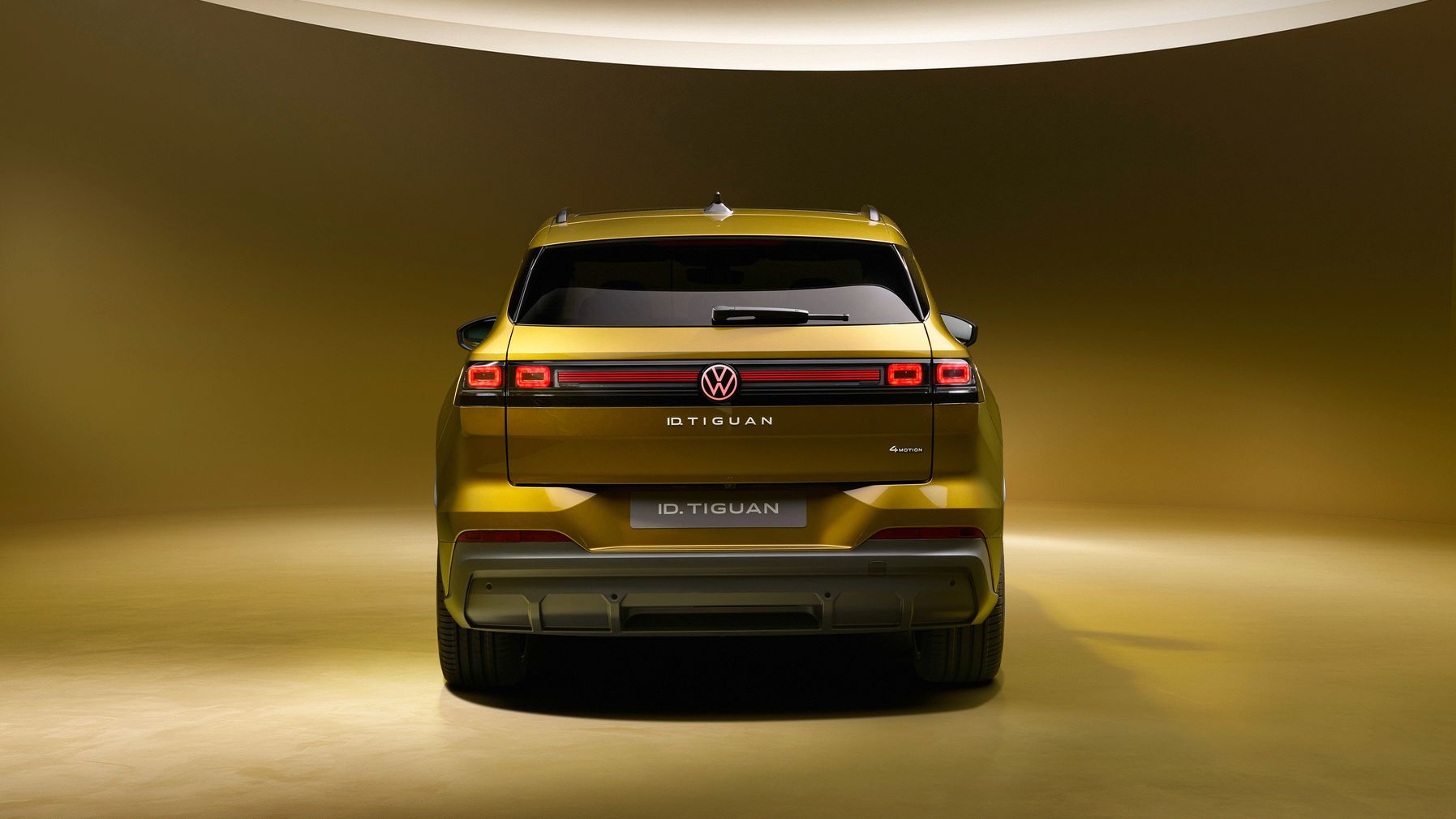 Rückansicht eines goldgelben VW ID. Tiguan in einem stilisierten Studio mit warm-goldener Beleuchtung. Das Fahrzeug wirkt futuristisch mit durchgehendem LED-Lichtband, markanten Rückleuchten und klaren, kantigen Linien. Im Fokus steht das Heckdesign mit dem mittig platzierten VW-Logo und dem Modellschriftzug.