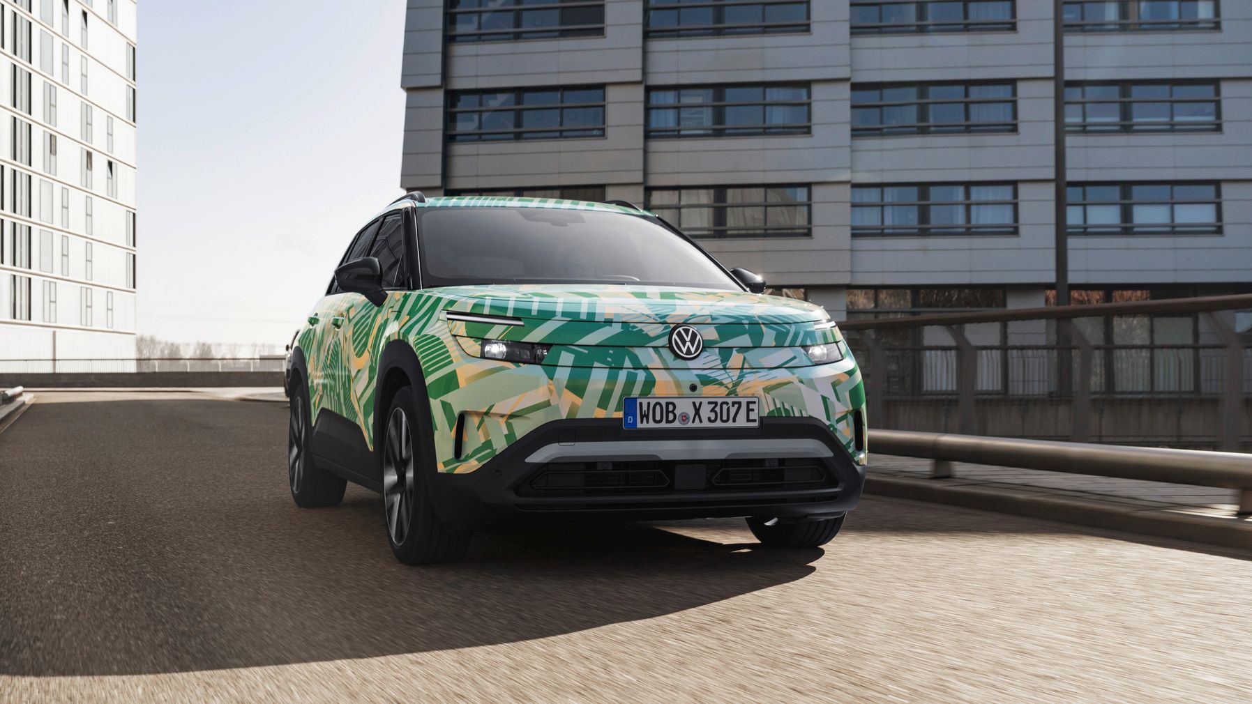 Ein ID. Cross von Volkswagen mit einem auffälligen grünen und gelben Camouflage-Design fährt auf einer städtischen Straße. Im Hintergrund sind moderne Gebäude zu sehen. Das Nummernschild des Autos beginnt mit "WOB".