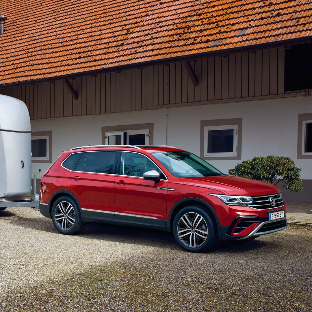 Roter Tiguan Allspace mit silbernem Pferdeanhänger steht auf einem Kieshof vor einem langgezogenen Wohn- oder Wirtschaftsgebäude mit roten Dachziegeln. Das Fahrzeug ist seitlich zu sehen, der Anhänger ist zweiachsig und geschlossen. Im Hintergrund befinden sich weiße Hauswände mit mehreren Fenstern und Türen.
