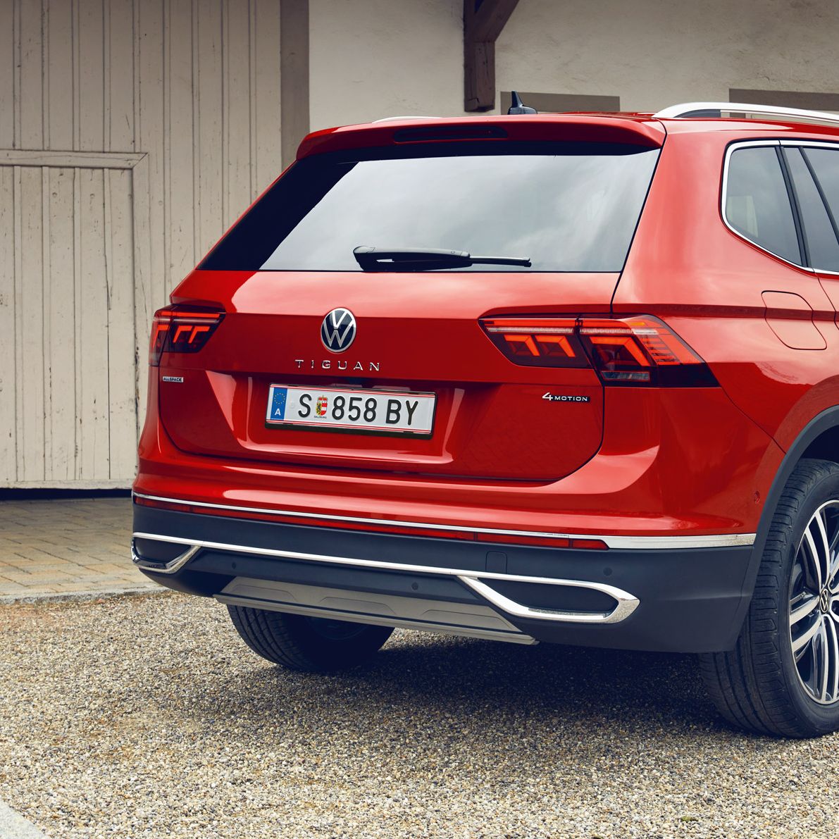 Roter VW Tiguan Allspace steht auf einem Kiesplatz vor einem hellen Gebäude mit Holztor. Das Auto ist aus schräger Heckansicht zu sehen, mit erkennbaren Rückleuchten, Modellschriftzug und österreichischem Kennzeichen.