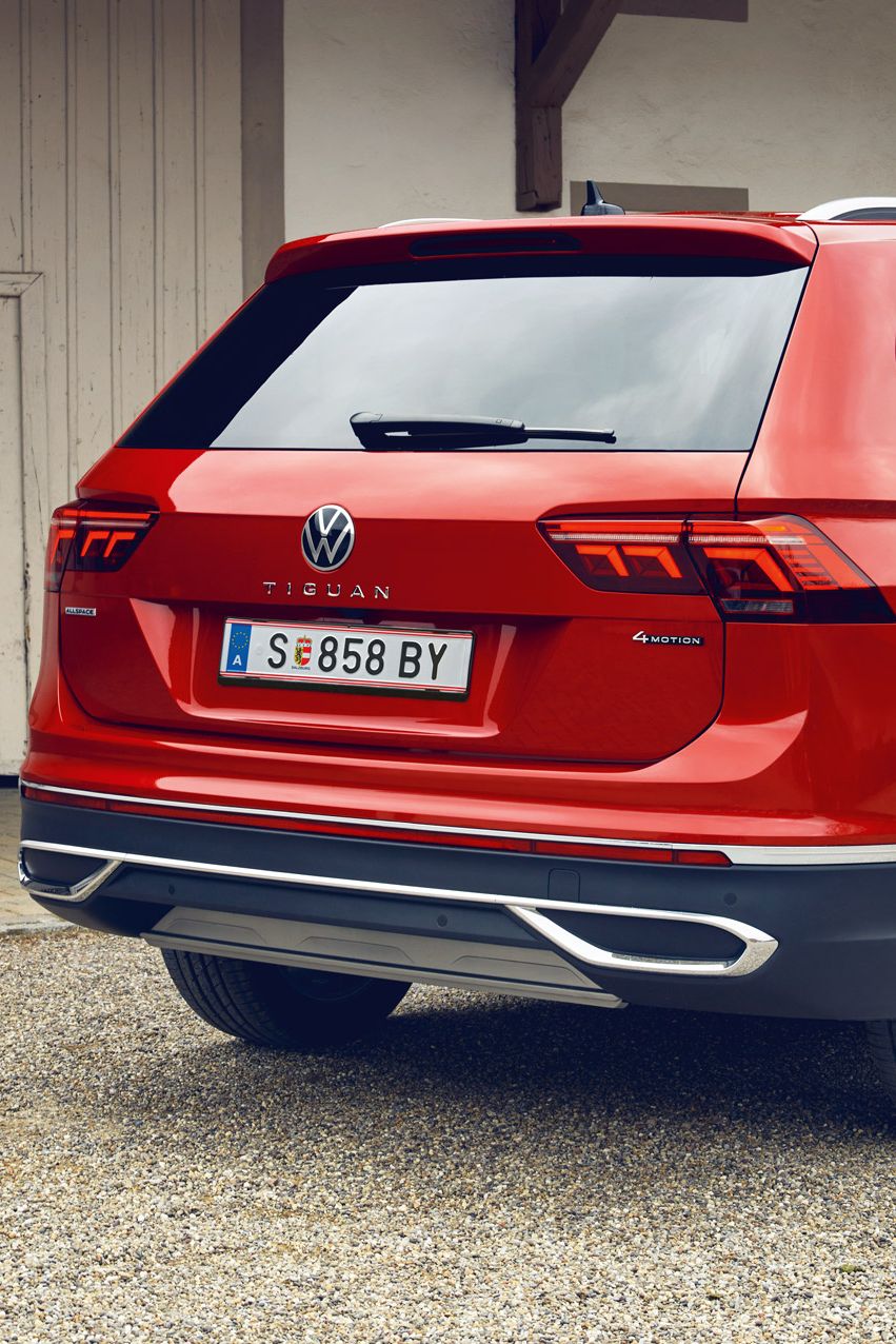 Roter VW Tiguan Allspace steht auf einem Kiesplatz vor einem hellen Gebäude mit Holztor. Das Auto ist aus schräger Heckansicht zu sehen, mit erkennbaren Rückleuchten, Modellschriftzug und österreichischem Kennzeichen.