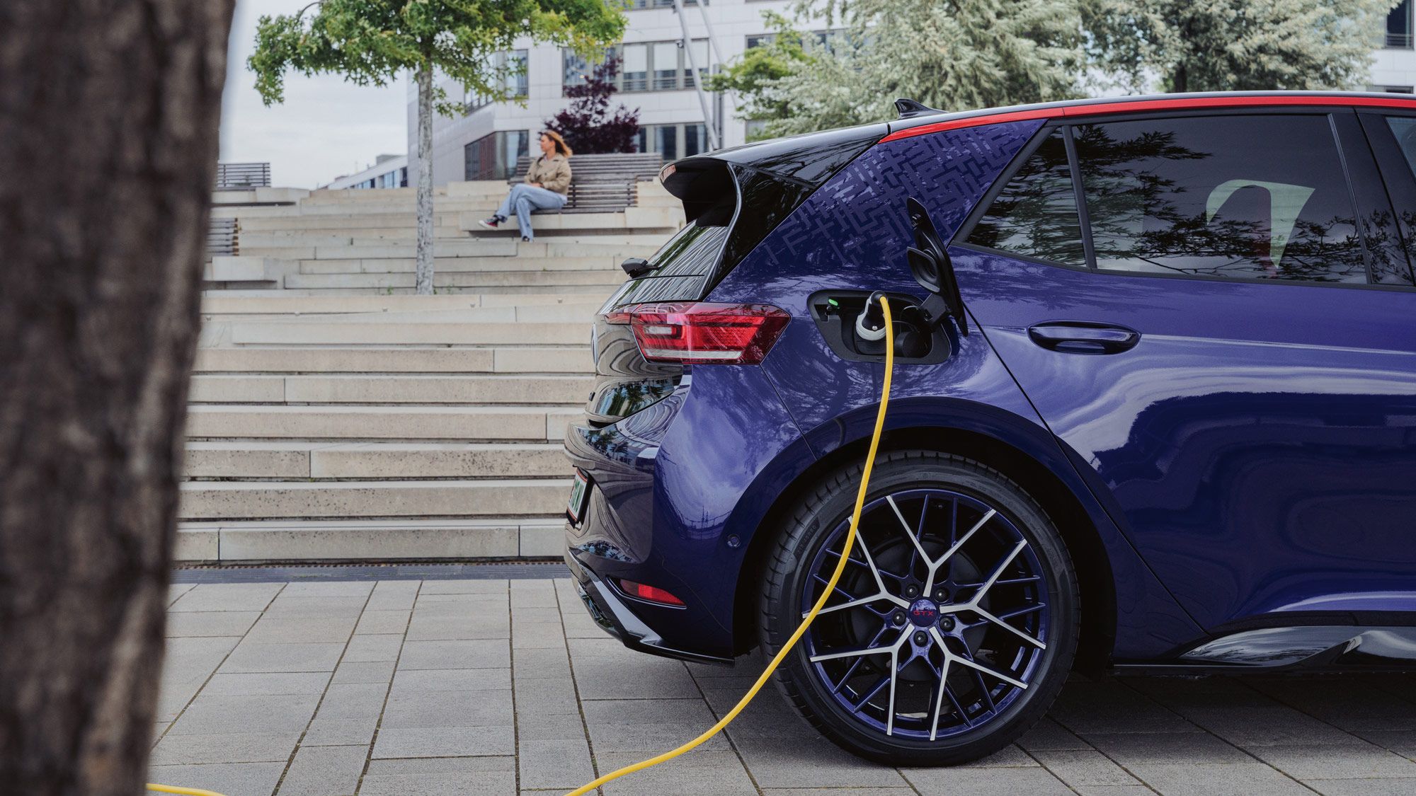 Ein Elektroauto wird auf einem Parkplatz mit einem gelben Kabel geladen. Im Hintergrund sitzt eine Person auf Treppenstufen. Um das Auto herum sind Bäume und moderne Gebäude.