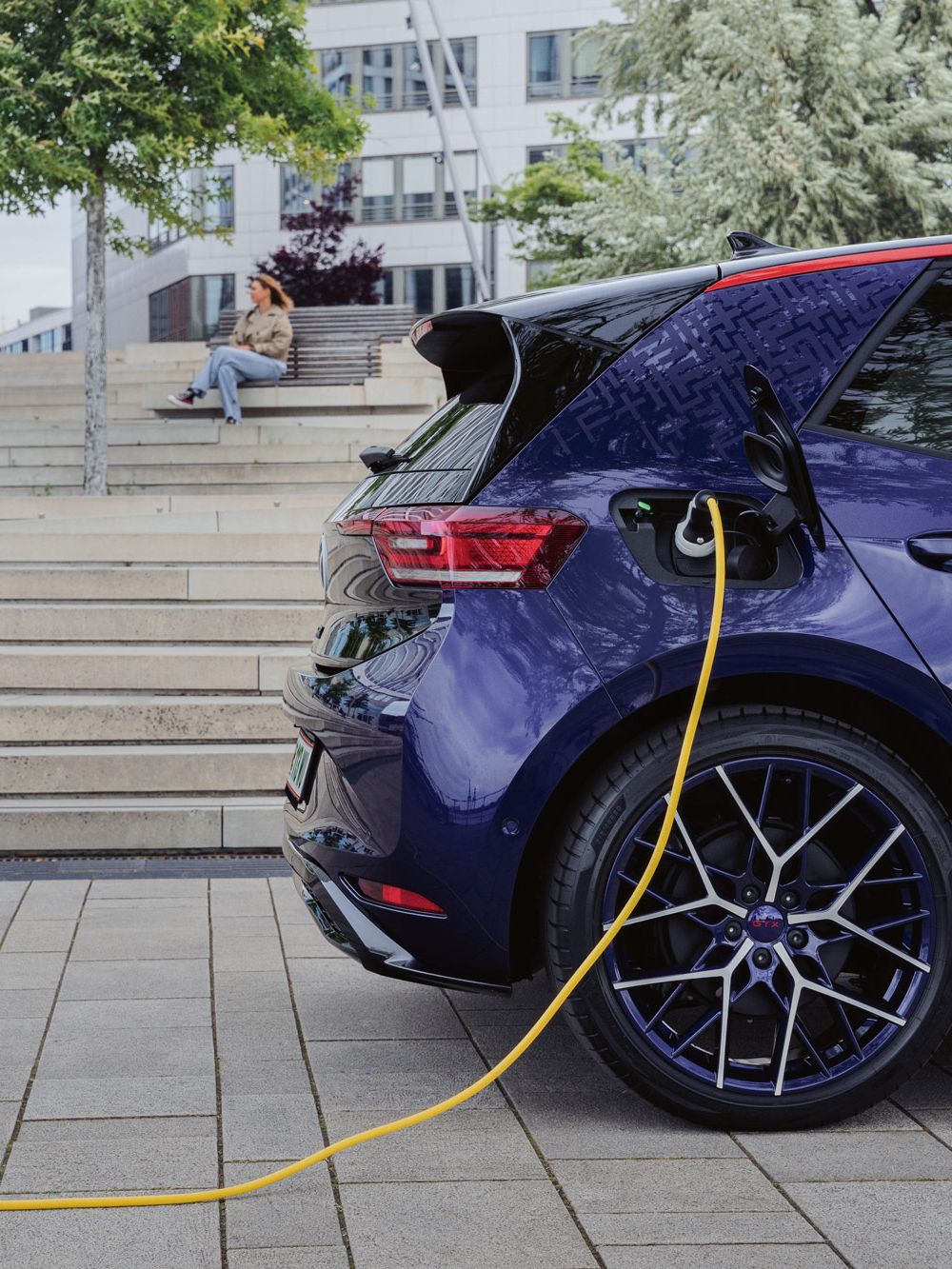 Ein Elektroauto wird auf einem Parkplatz mit einem gelben Kabel geladen. Im Hintergrund sitzt eine Person auf Treppenstufen. Um das Auto herum sind Bäume und moderne Gebäude.