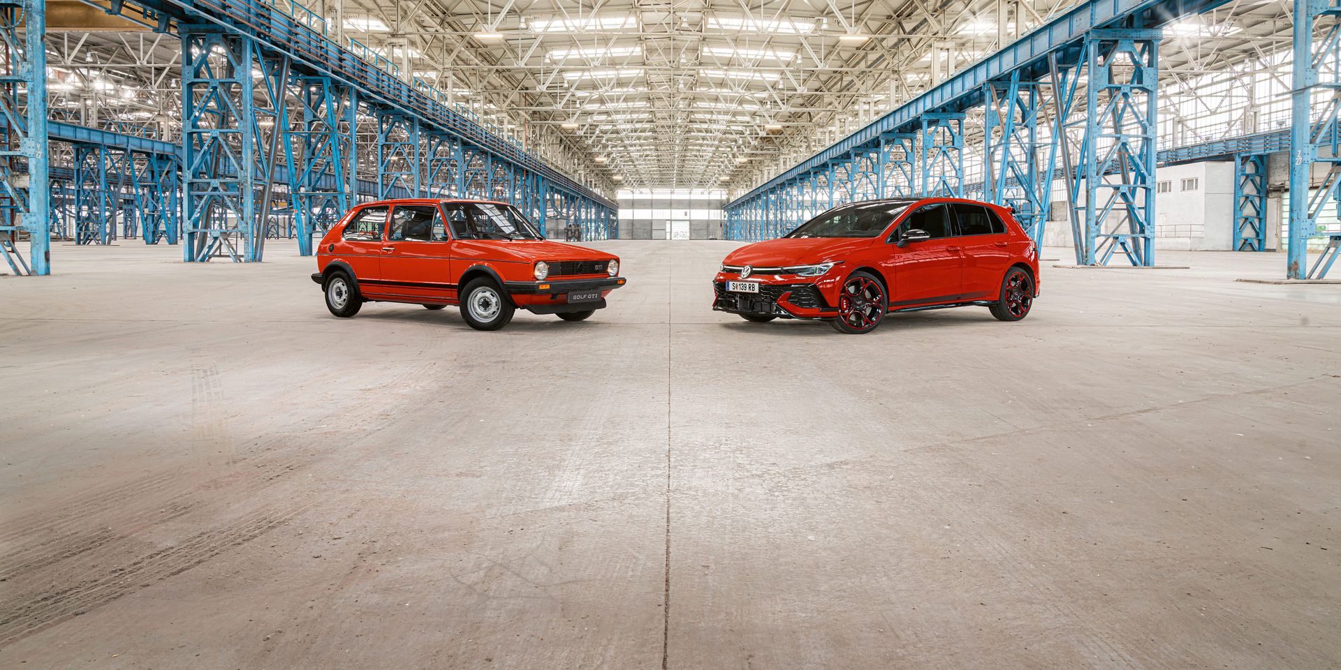 Zwei rote Golf GTI, das erste und das neuste Modell, stehen in einer großen Industriehalle mit vielen blauen Stahlträgern und einem hohen, hellen Dach.
