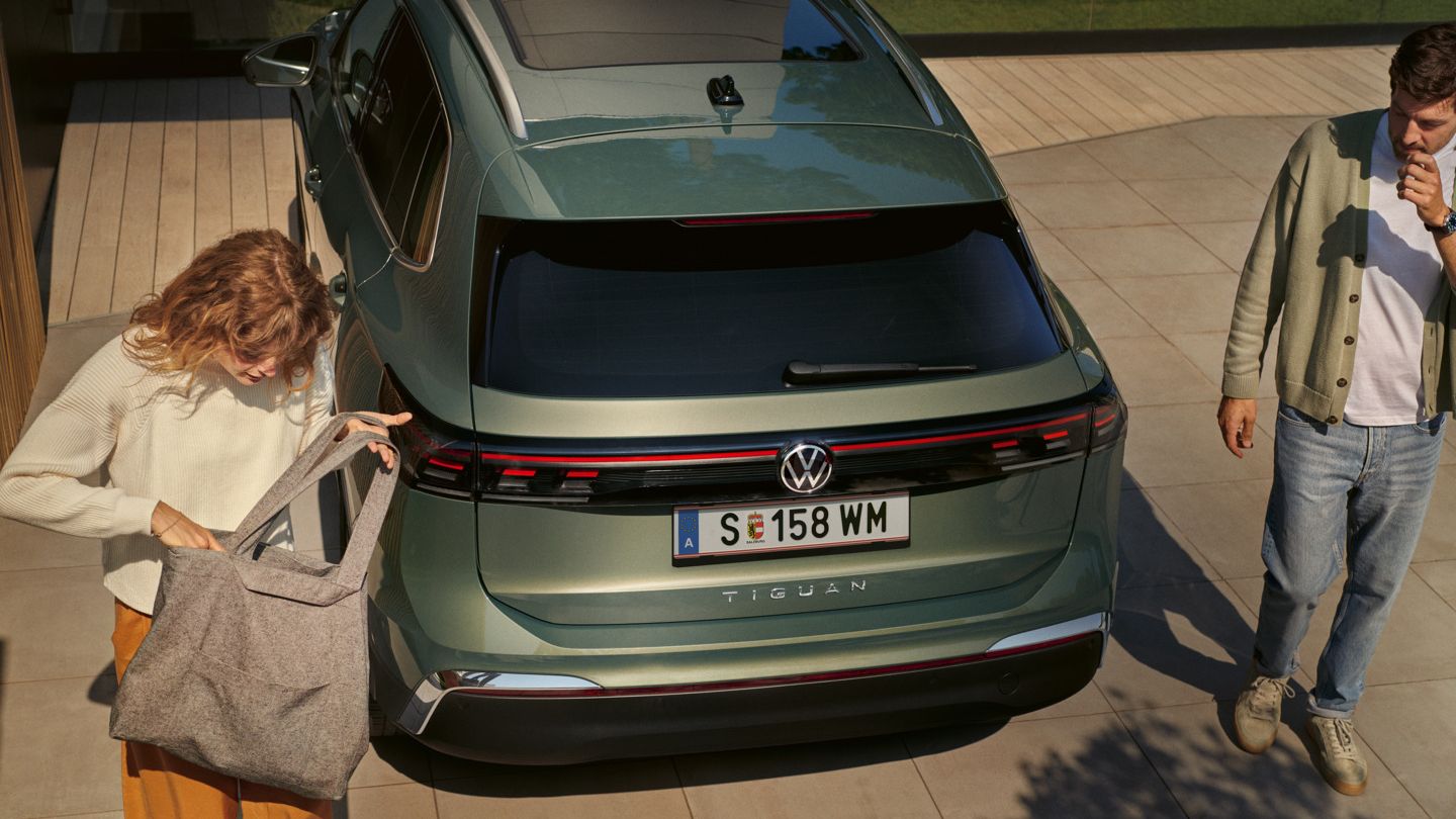 Ein Paar steht neben einem grünen VW Tiguan. Die Frau packt Gegenstände in eine Tasche. Das Fahrzeug ist auf einer gepflasterten Einfahrt geparkt.