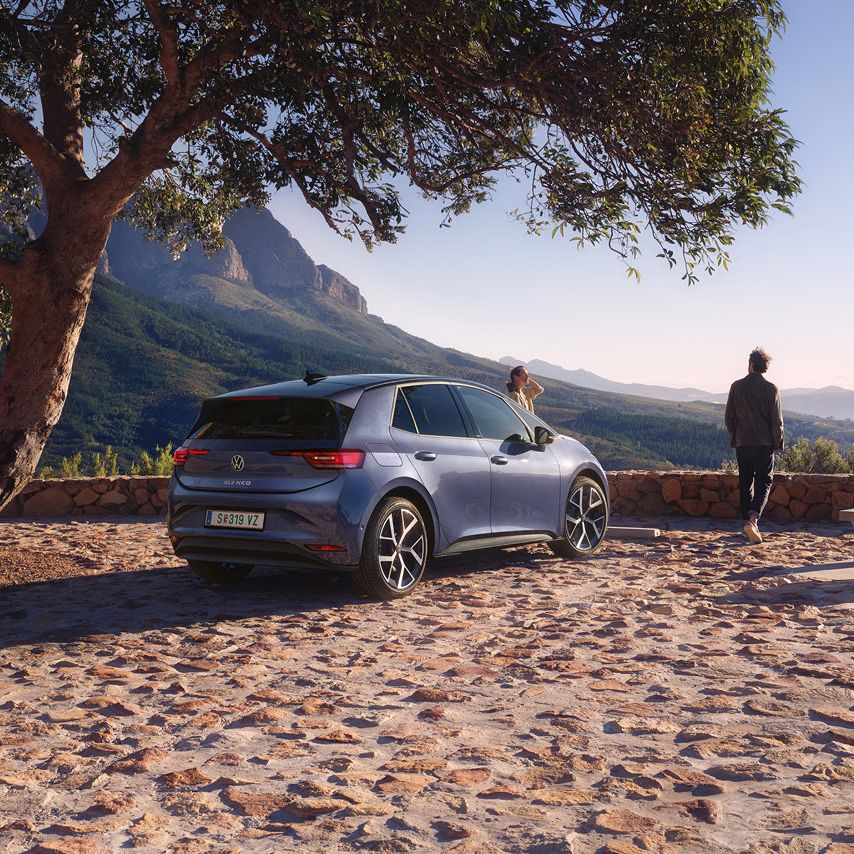Auf einem Parkplatz in den Bergen parkt ein blauer VW ID.3 Neo, eine Frau und ein Mann genießen die Aussicht