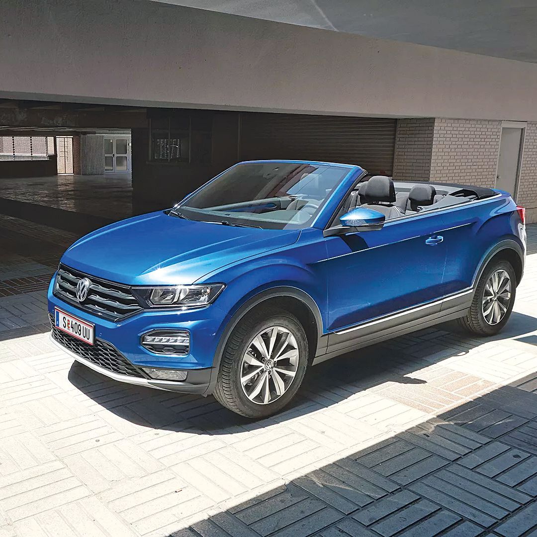 Ein blaues Volkswagen Cabriolet steht in einem sonnigen, überdachten Bereich. Es hat das Verdeck geöffnet. Die Umgebung ist modern und urban.