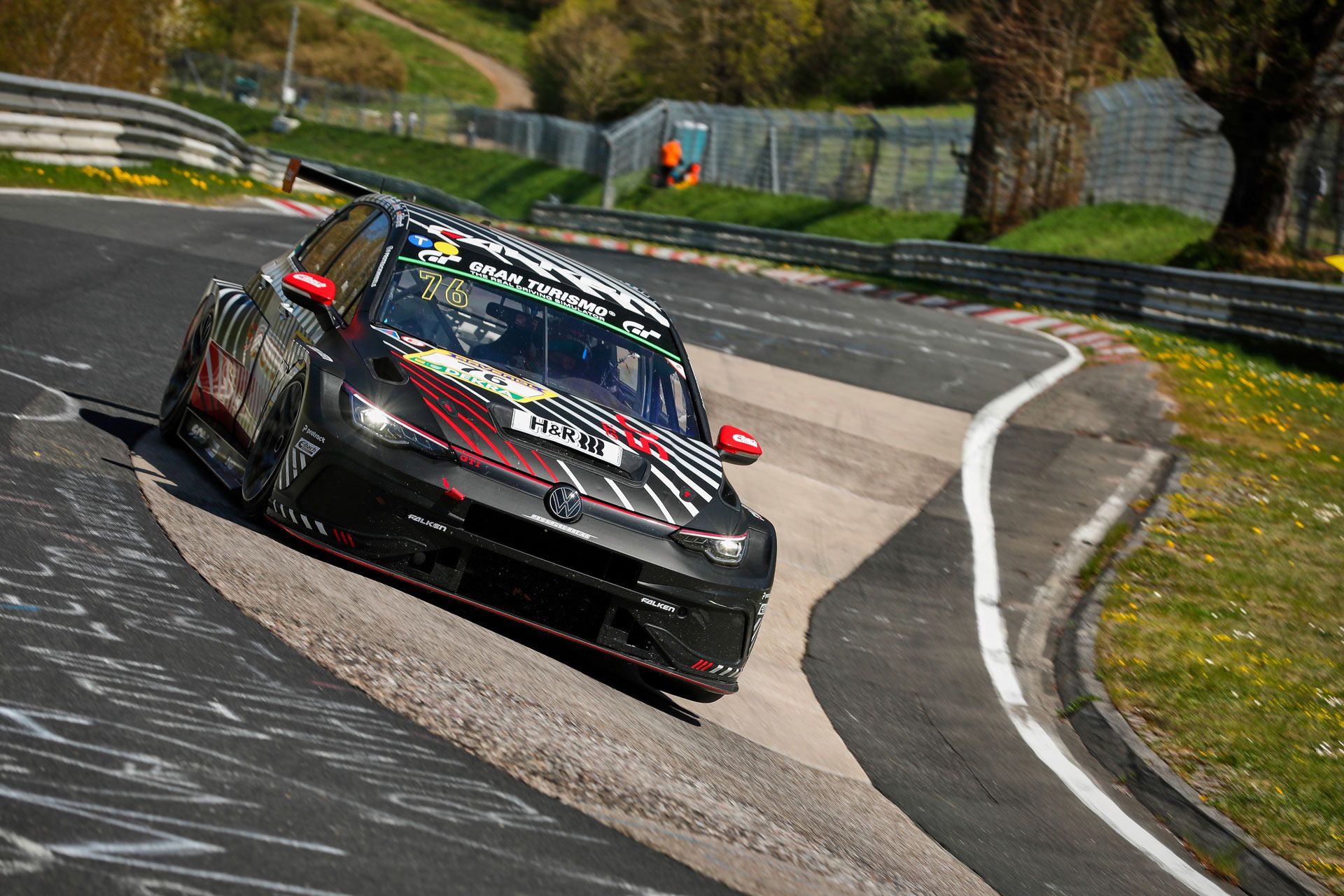 Ein Golf GTI Clubsport fährt eine Kurve auf einer Rennstrecke. Das Auto hat eine schwarz-rote Lackierung und fährt durch eine grüner Umgebung. Im Hintergrund sind Streckenbegrenzungen und Zuschauerbereiche zu sehen.