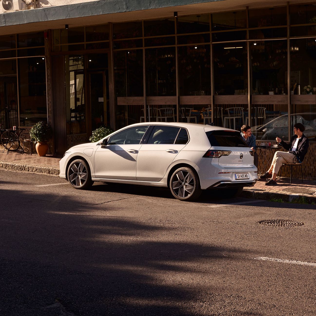 Zwei Männer sitzen vor einem Cafe und unterhalten sich. Vor ihnen ein neuer Golf 8.