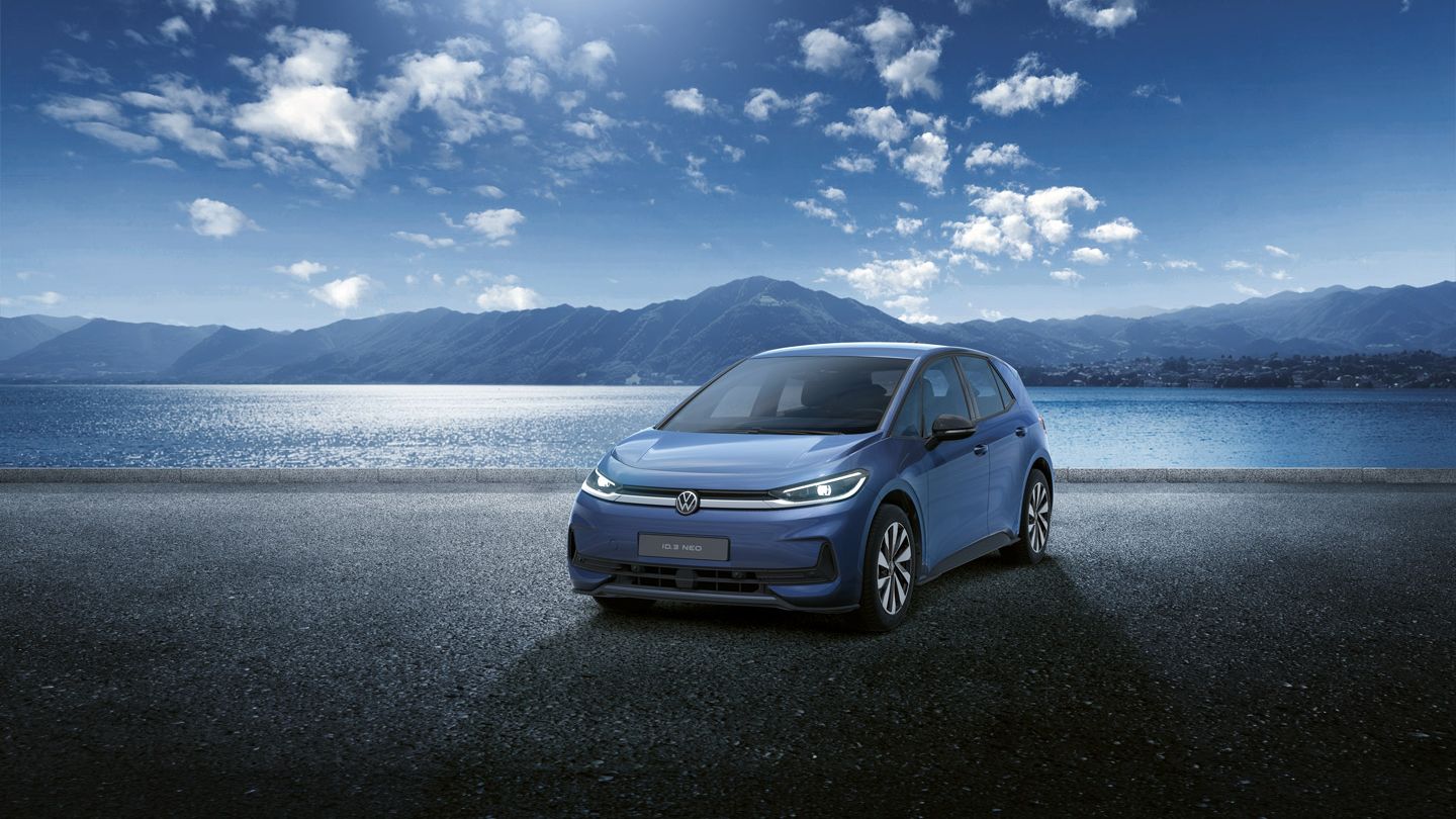 Blauer VW ID.3 Neo steht auf einer asphaltierten Fläche vor einem weiten See mit Bergkette im Hintergrund. Der Himmel ist klar mit einigen Wolken, die Sonne scheint von oben. Die Szene wirkt ruhig und modern, mit Fokus auf dem Elektroauto in der Mitte.