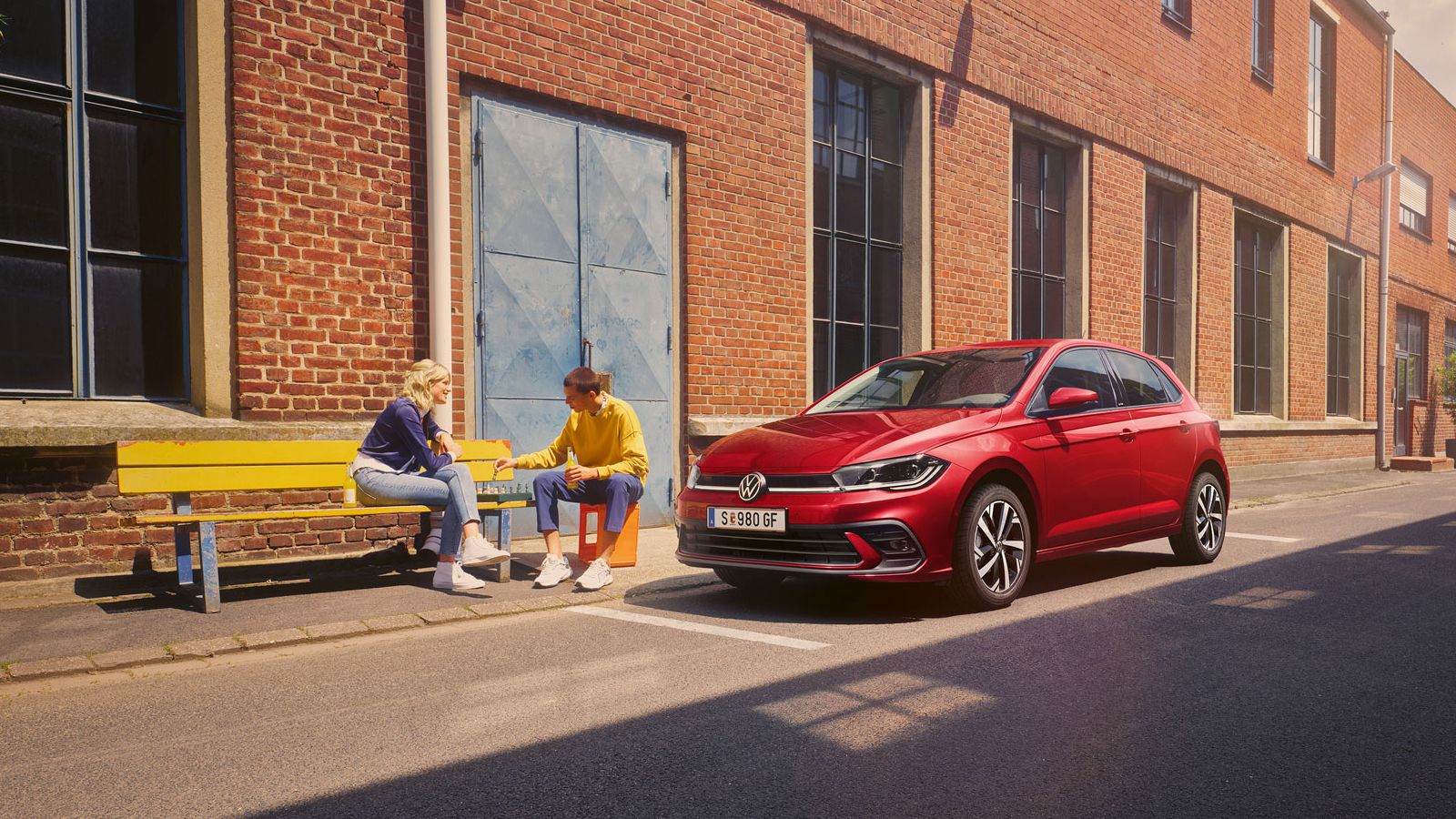 Zwei Personen spielen Schach vor einem roten VW Polo Style, der vor einem Fabrikgebäude parkt.