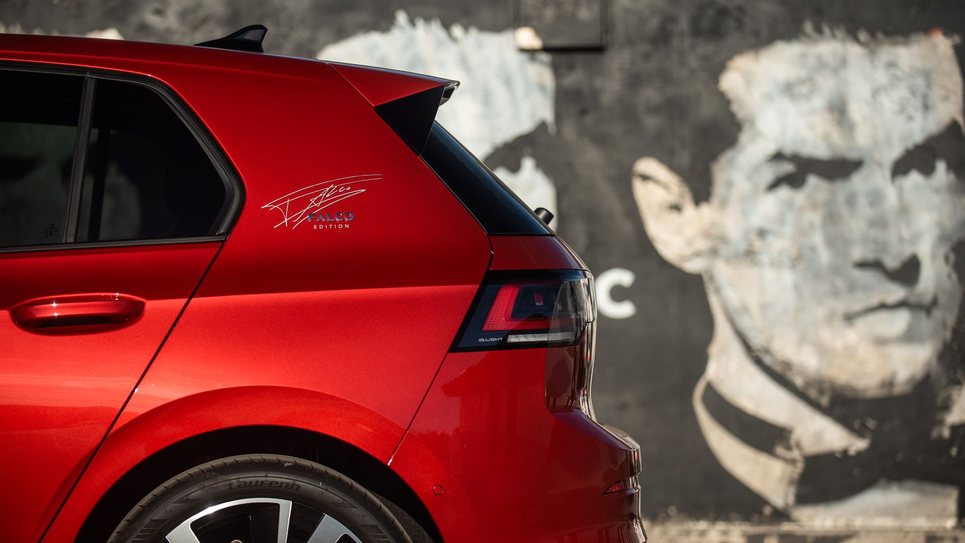Rückansicht eines roten Kompaktwagens, bei dem vor allem das linke Heck und das Hinterrad im Vordergrund stehen. Im Hintergrund ist eine dunkle Wand mit verblasster, teils abblätternder Streetart-Grafik zu sehen. Rechts im Bild ist ein unscharfer, neutraler Bereich ohne erkennbare Details.