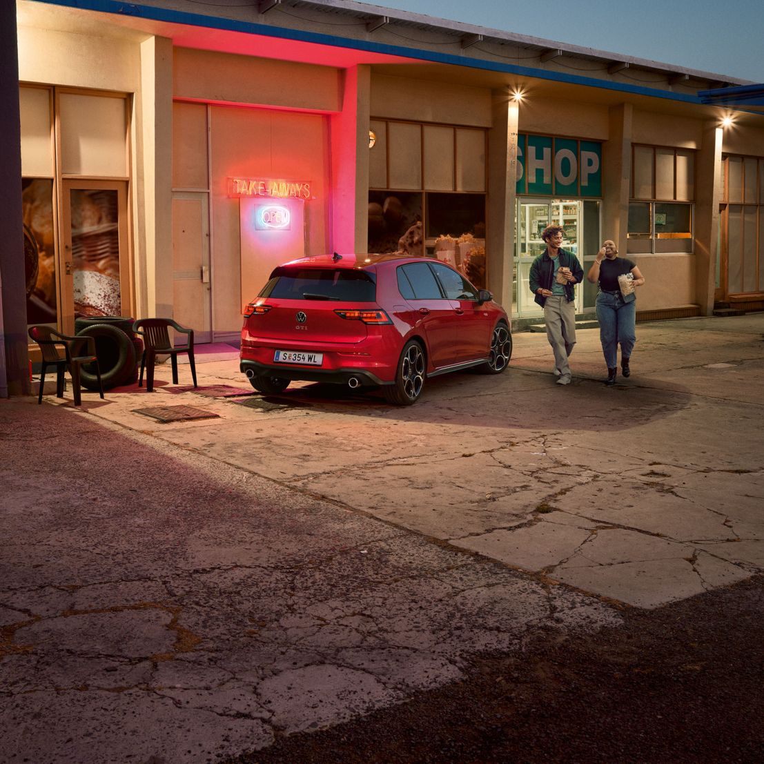 Roter VW Golf GTI steht abends vor einem Supermarkt, zwei junge Leute gehen mit Einkäufen daran vorbei.