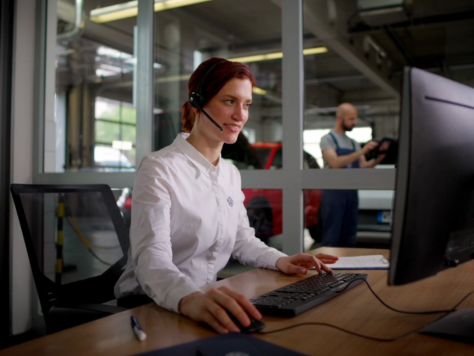 Eine Frau mit Kopfhörern arbeitet an einem Computer. Im Hintergrund ist eine Werkstatt mit einem Mechaniker zu sehen.