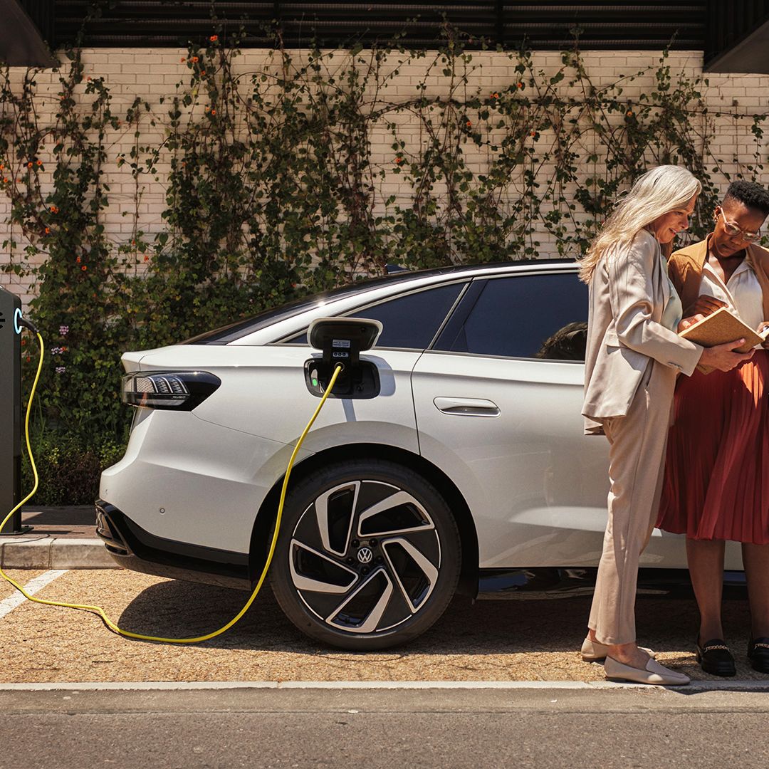 Zwei Frauen stehen mit einem Buch in der Hand neben einem weißen VW ID., der an einer öffentlichen Ladesäule lädt.