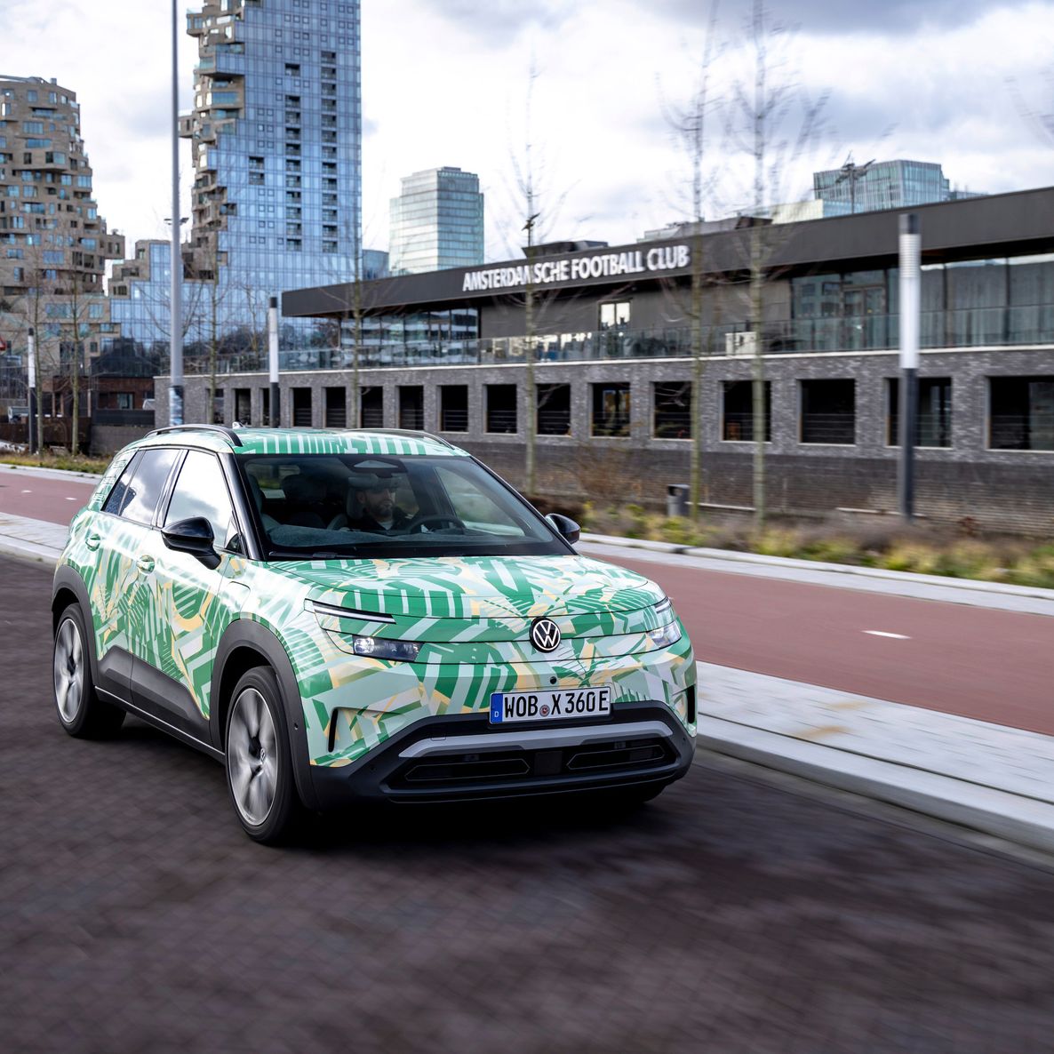 Ein Elektroauto fährt auf einer städtischen Straße an modernen Gebäuden vorbei. Das Fahrzeug hat ein auffälliges Design mit grünen und gelben Mustern. Im Hintergrund ist ein Gebäude mit der Aufschrift "Mitsubishi Electric Football Club" zu sehen.