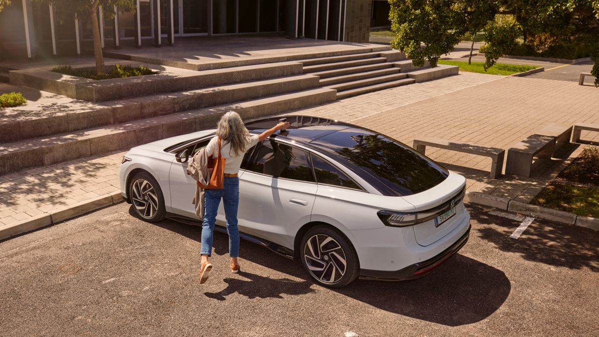 Eine Frau nimmt einen Becher vom Dach eines VW ID.7, der vor einem Bürogebäude parkt.