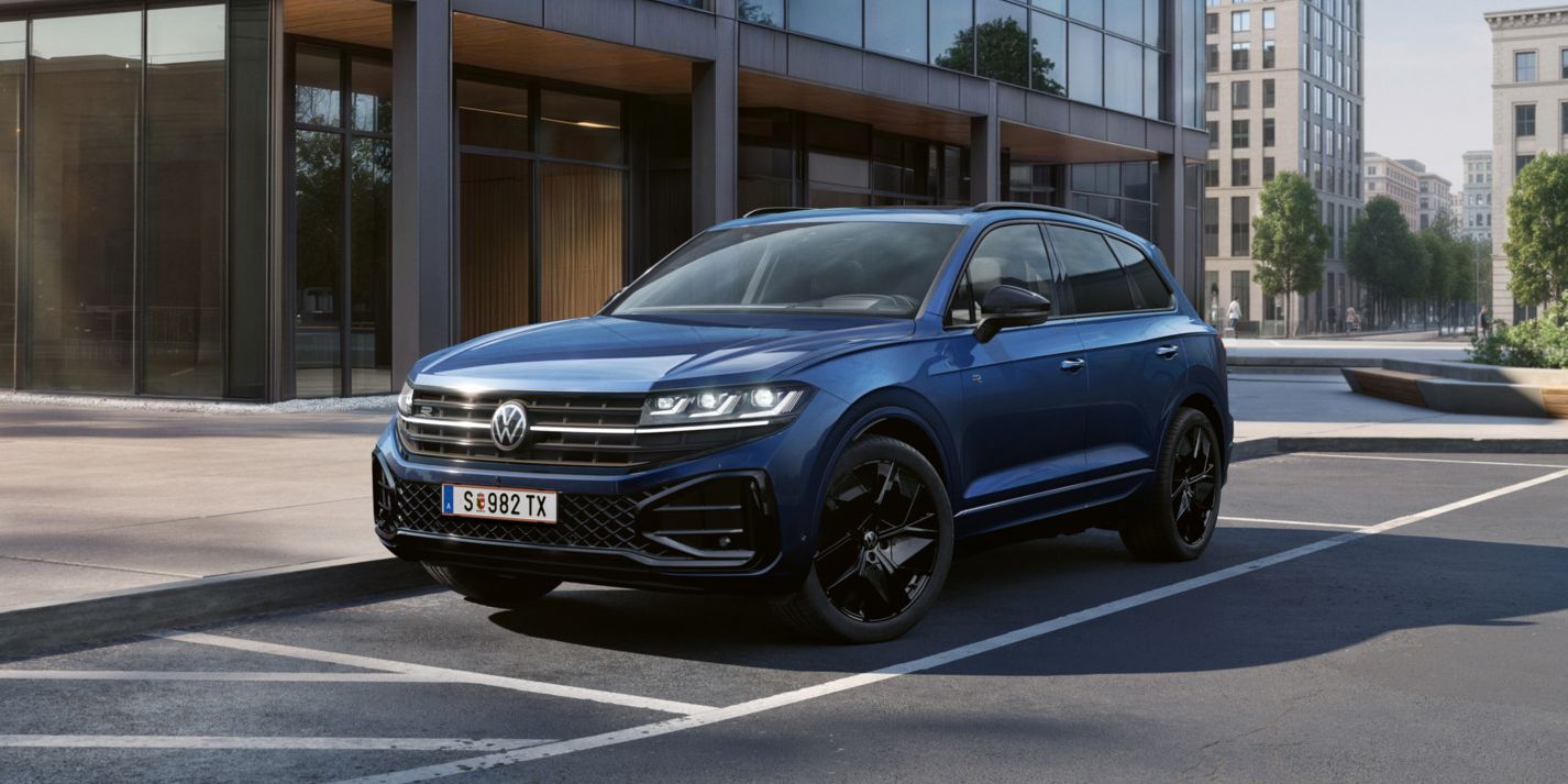 Ein blauer VW Touareg Final Edition steht vor einem Bürogebäude mit Glasfassade - Ansicht von schräg vorne.