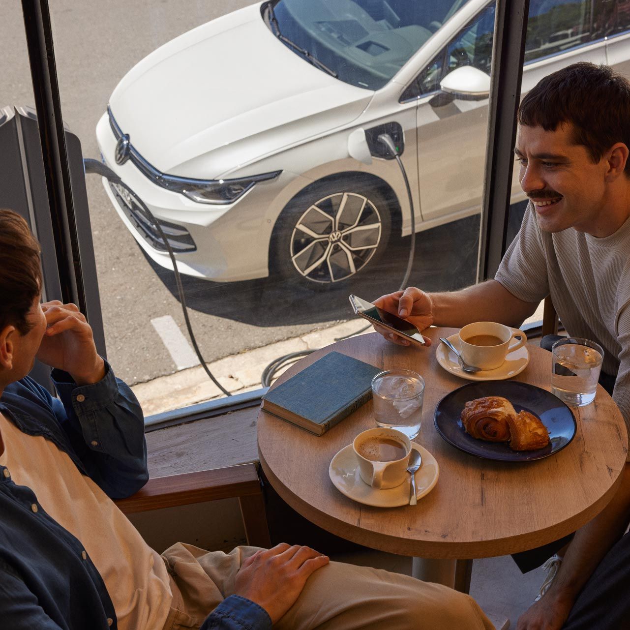 Zwei Männer sitzen in Café und unterhalten sich, durch das Fenster sieht man einen VW Golf eHybrid der geladen wird