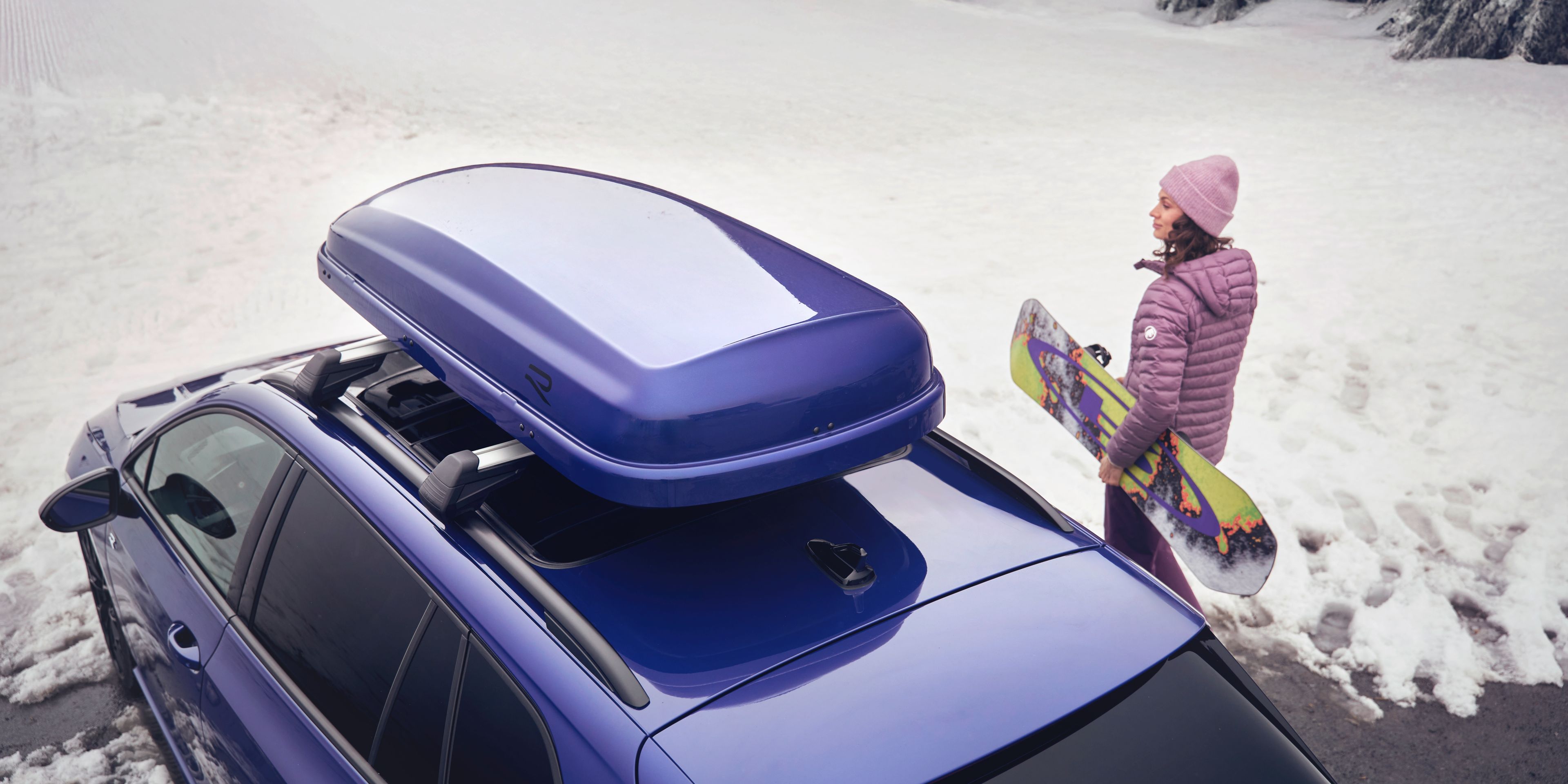 Blick von oben auf eine Frau mit Snowboard neben einem blauen Volkswagen  mit blauer Dachbox in einer verschneiten Landschaft.