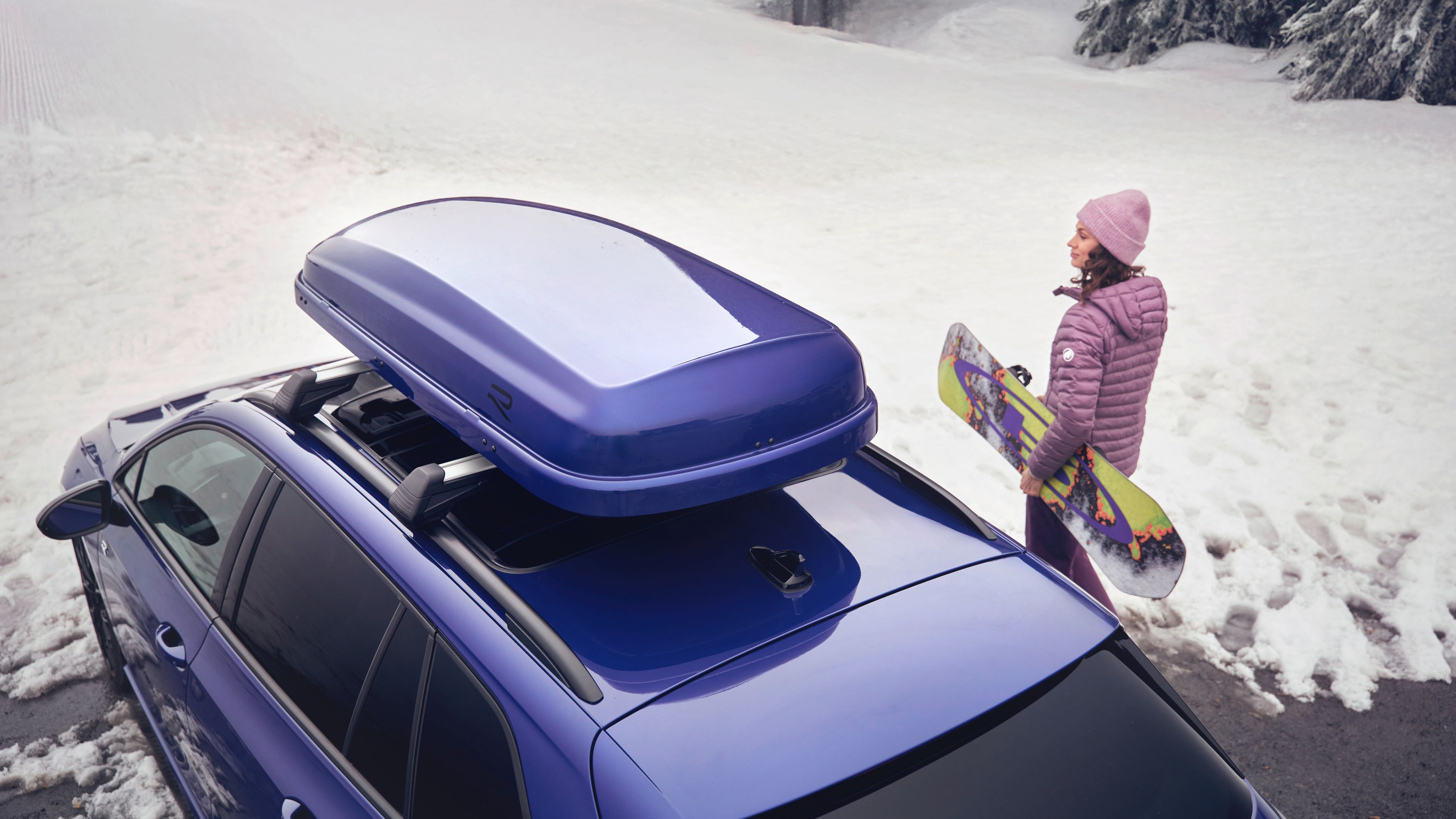 Blick von oben auf eine Frau mit Snowboard neben einem blauen Volkswagen  mit blauer Dachbox in einer verschneiten Landschaft.