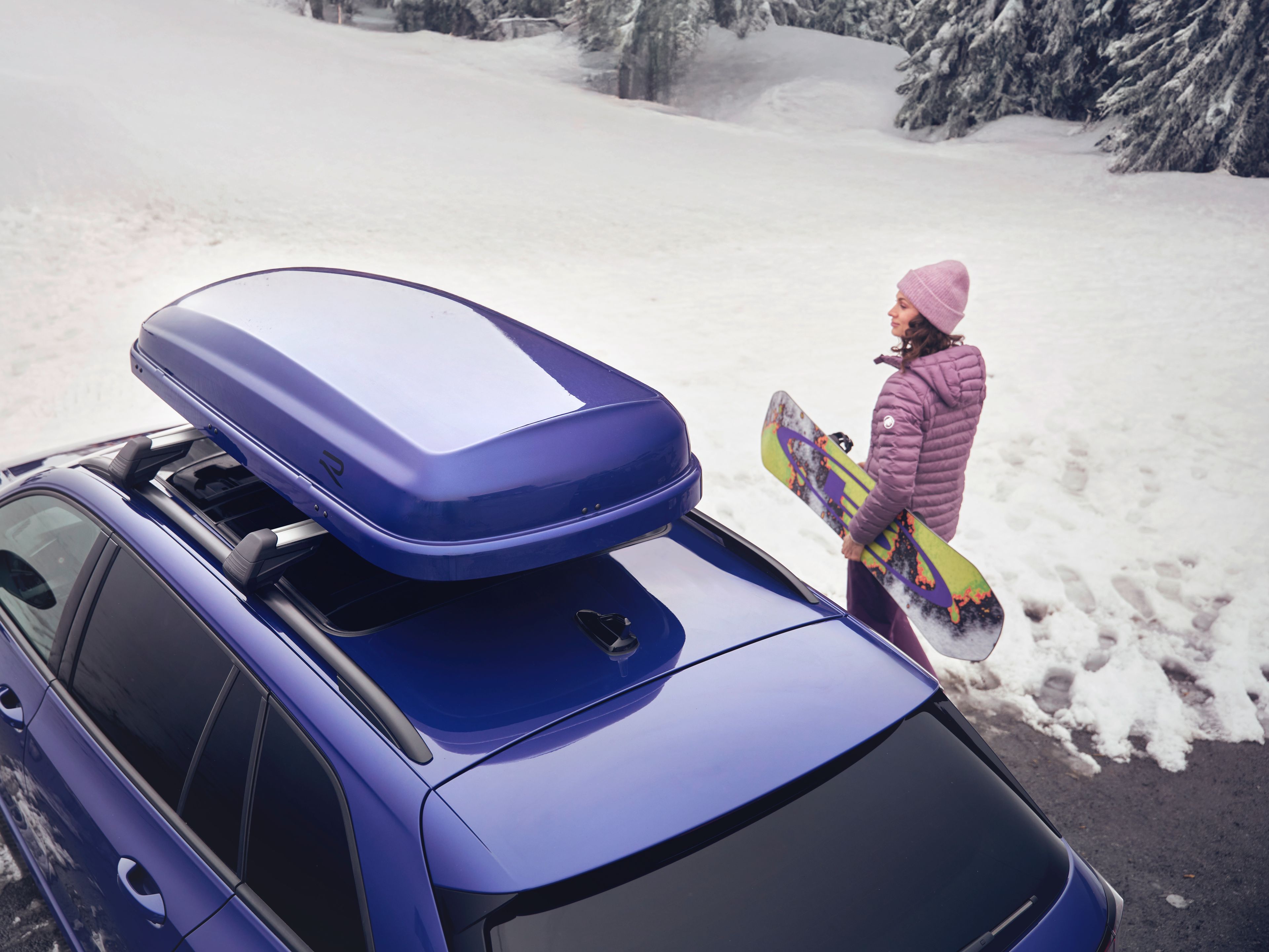 Blick von oben auf eine Frau mit Snowboard neben einem blauen Volkswagen  mit blauer Dachbox in einer verschneiten Landschaft.