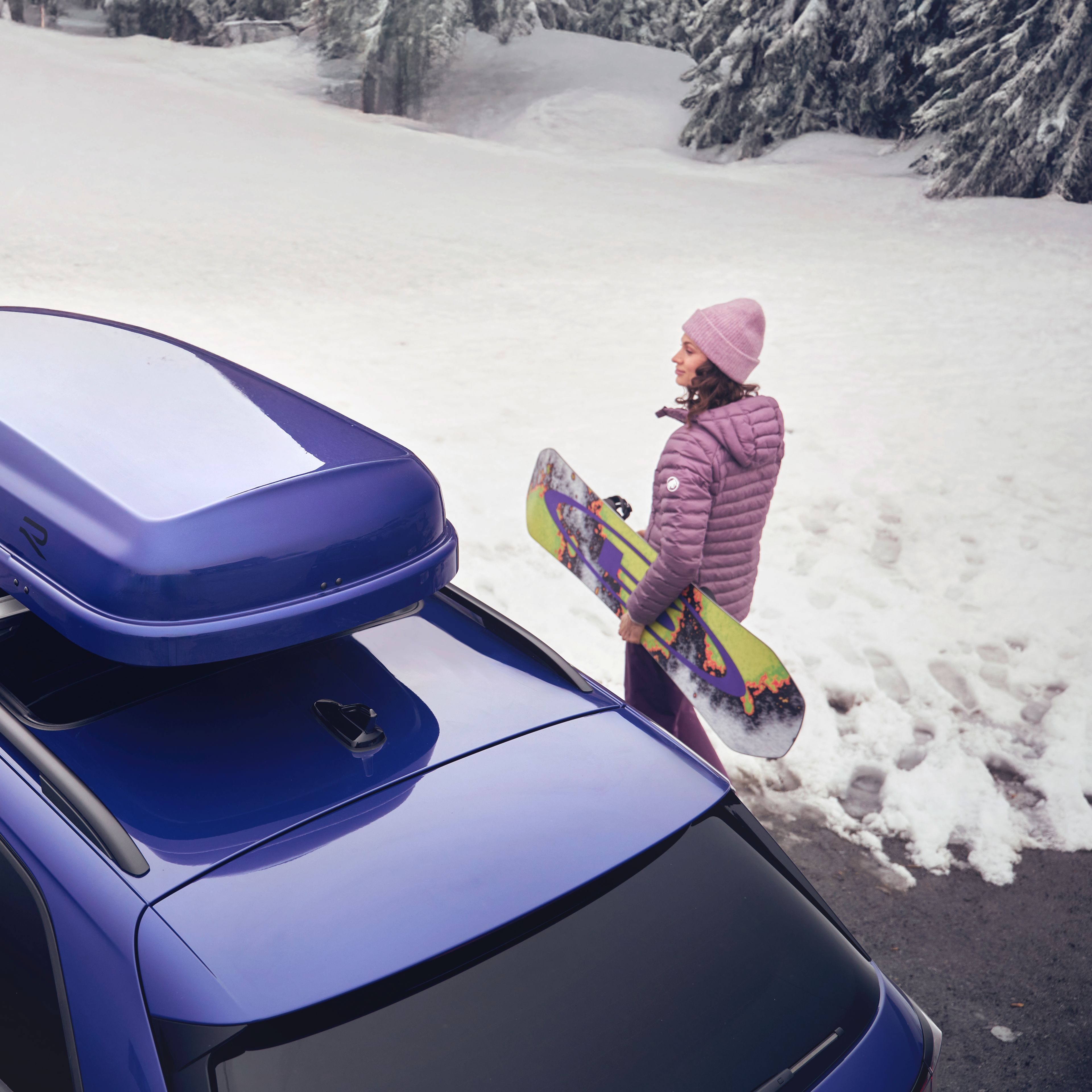 Blick von oben auf eine Frau mit Snowboard neben einem blauen Volkswagen  mit blauer Dachbox in einer verschneiten Landschaft.
