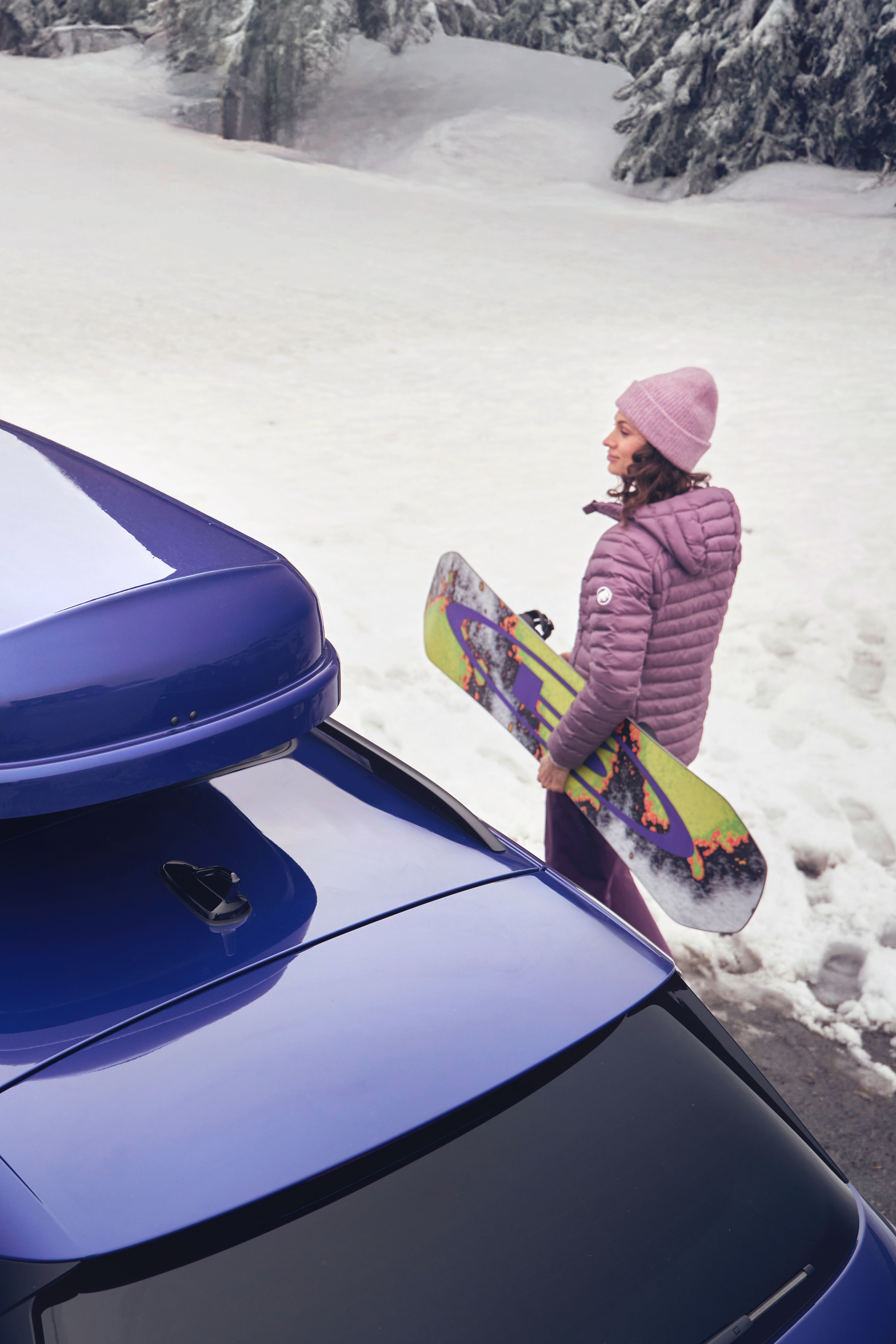 Blick von oben auf eine Frau mit Snowboard neben einem blauen Volkswagen  mit blauer Dachbox in einer verschneiten Landschaft.