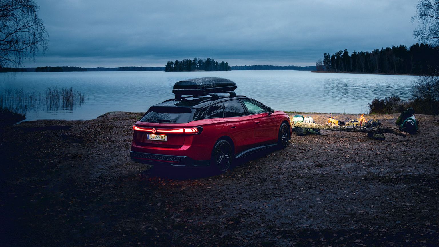 Roter VW ID.7 GTX Tourer mit Dachbox parkt an einem See in der Dämmerung.