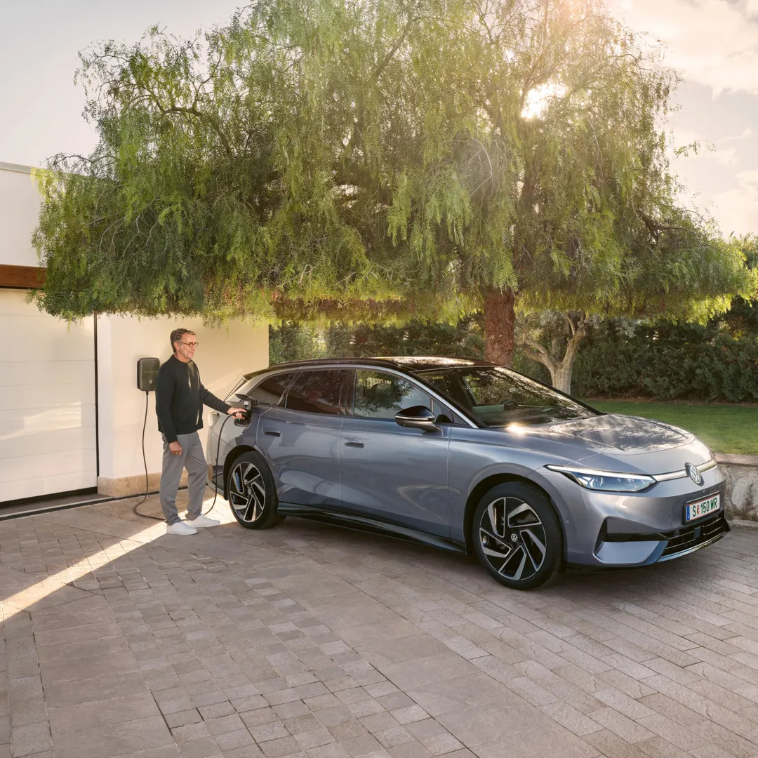 Ein Mann steht neben einem blauen VW ID.7 Tourer, der vor einer Garage an einer Wallbox lädt.