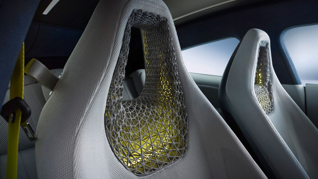 Innenraum eines modernen Sportwagens mit zwei futuristischen Schalensitzen, deren Rückenlehnen aus einem durchlässigen, gitterartigen 3D-Muster bestehen. Durch das Netzgewebe scheint gelbes Licht hindurch, was den technischen, hochwertigen Eindruck verstärkt. Links ist ein gelber Sicherheitsgurt zu erkennen.