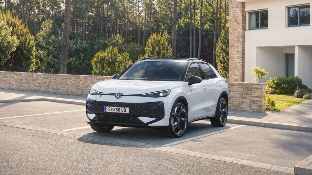 Ein weißer VW T-Roc Hybrid steht auf einem Parkplatz vor einem Wohnhaus am Wald, Blick auf die Front.