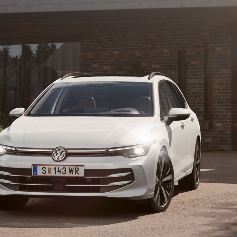 Ein weißer VW Golf Variant parkt in der Auffahrt eines Bungalows mit aktivem Tagfahrlicht.