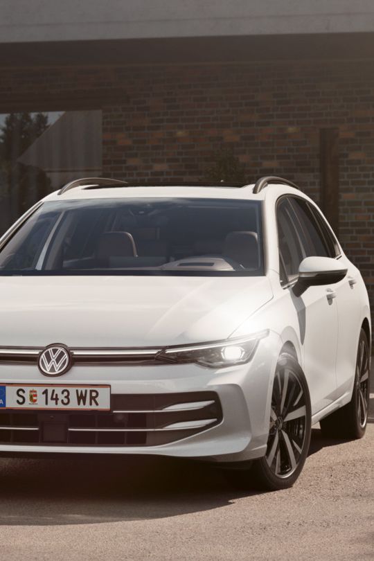 Ein weißer VW Golf Variant parkt in der Auffahrt eines Bungalows mit aktivem Tagfahrlicht.