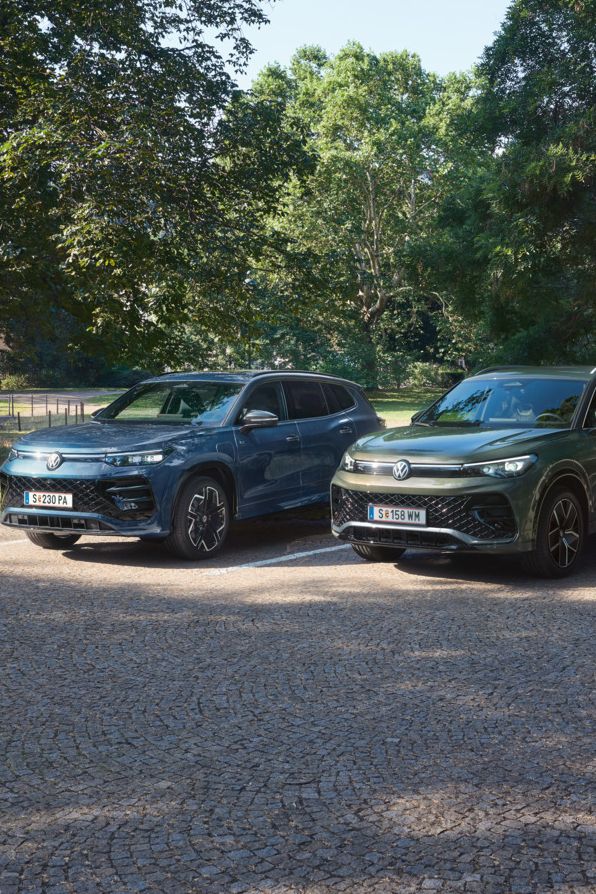 Blauer Tiguan R-Line Plug-In-Hybrid und grüner Tayron R-Line Plug-In- Hybrid parken nebeneinander im schattigen Parkbereich.