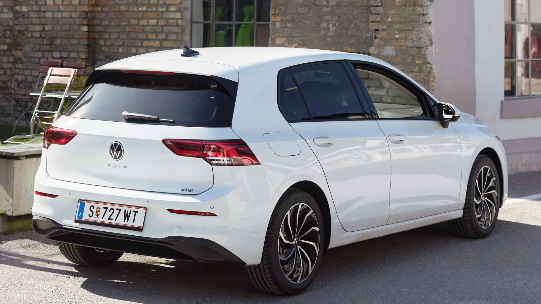 Ein weißer Volkswagen Golf eTSI steht vor einem Gebäude aus Ziegelsteinen. Neben dem Auto stehen einige bunte Stühle. Das Auto hat ein österreichisches Kennzeichen.
