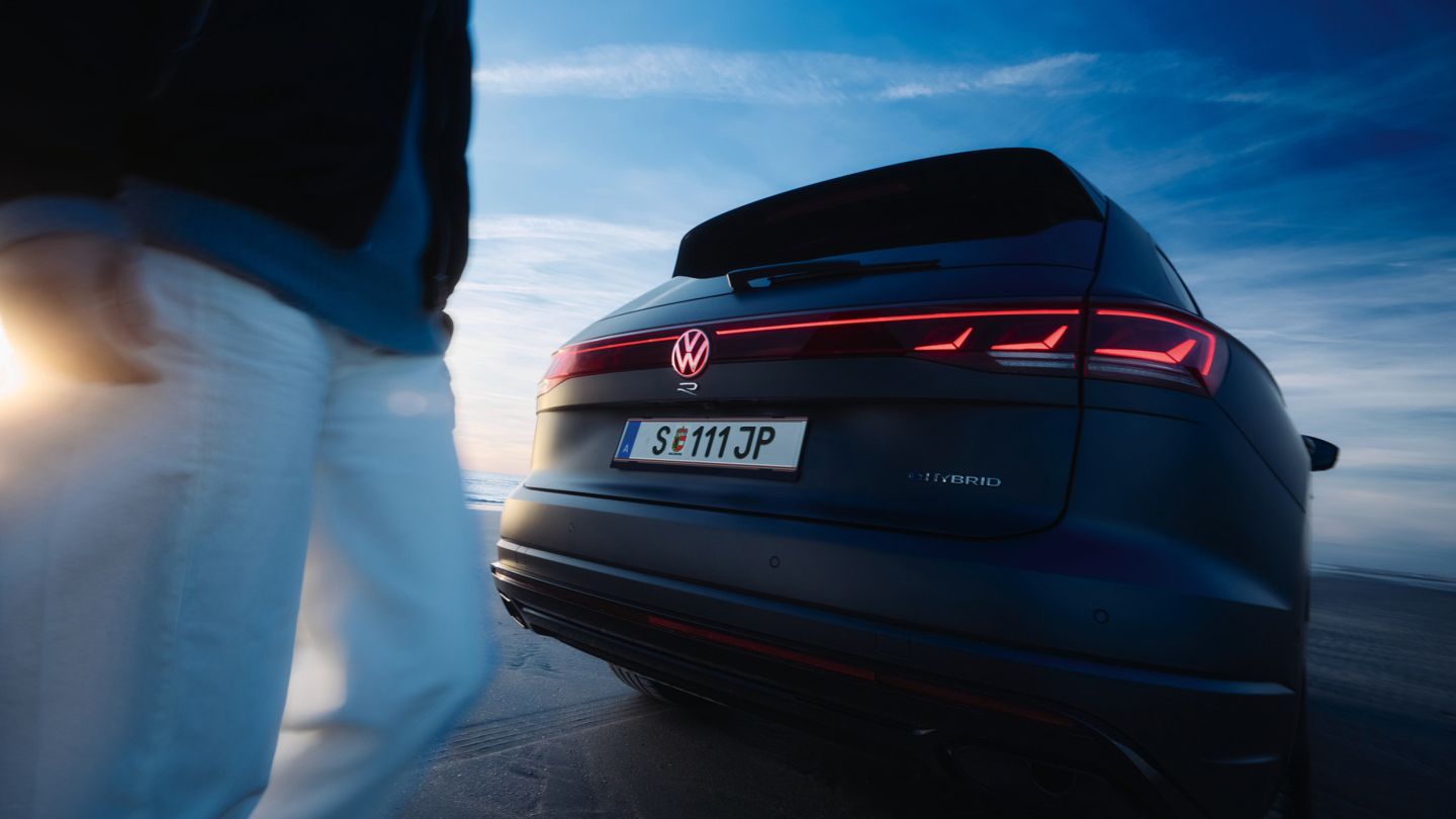 Ein Touareg R steht auf einem sandigen Untergrund. Die Rücklichter leuchten rot und ein Mensch befindet sich auf der linken Bildseite. Im Hintergrund sind blauer Himmel und Wolken zu sehen.