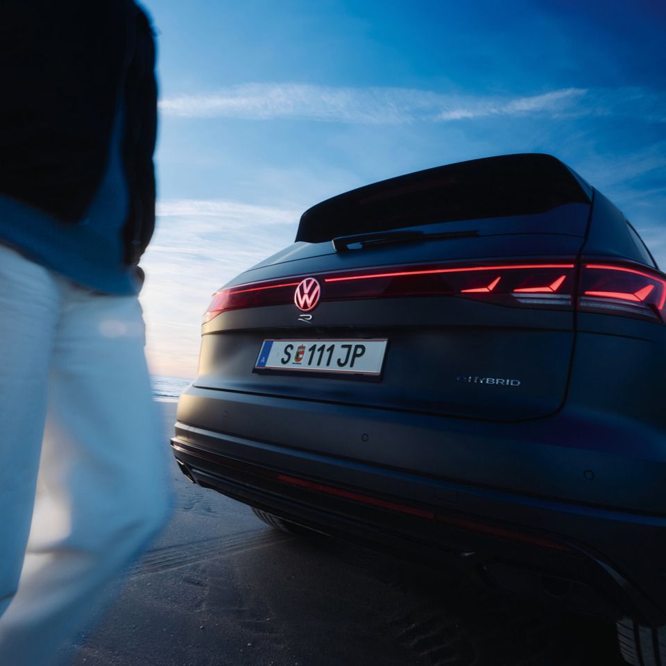 Ein Touareg R steht auf einem sandigen Untergrund. Die Rücklichter leuchten rot und ein Mensch befindet sich auf der linken Bildseite. Im Hintergrund sind blauer Himmel und Wolken zu sehen.