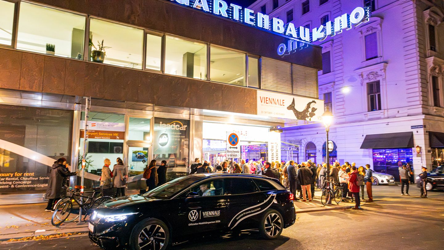 Abendliche Straßenszene vor dem beleuchteten „Gartenbaukino“ in Wien mit vielen Menschen, die vor dem Eingang anstehen oder sich unterhalten. Ein schwarzes Auto mit „Viennale“- und VW‑Logo fährt im Vordergrund an der Menschenmenge vorbei. Die Umgebung ist herbstlich mit Laub auf dem Boden und warmem Kunstlicht, das die Szene stimmungsvoll ausleuchtet.