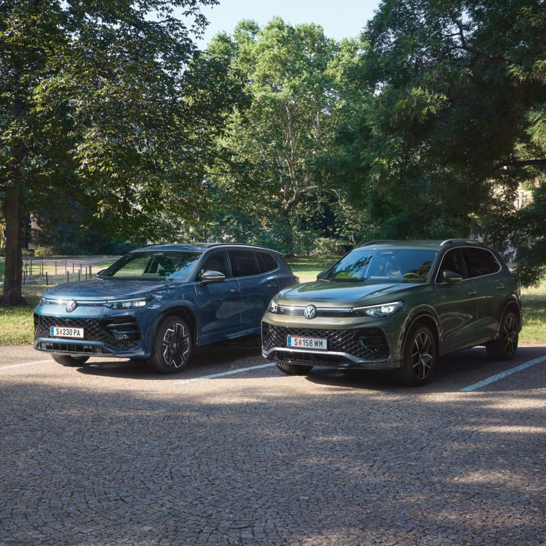 Blauer Tiguan R-Line Plug-In-Hybrid und grüner Tayron R-Line Plug-In- Hybrid parken nebeneinander im schattigen Parkbereich.