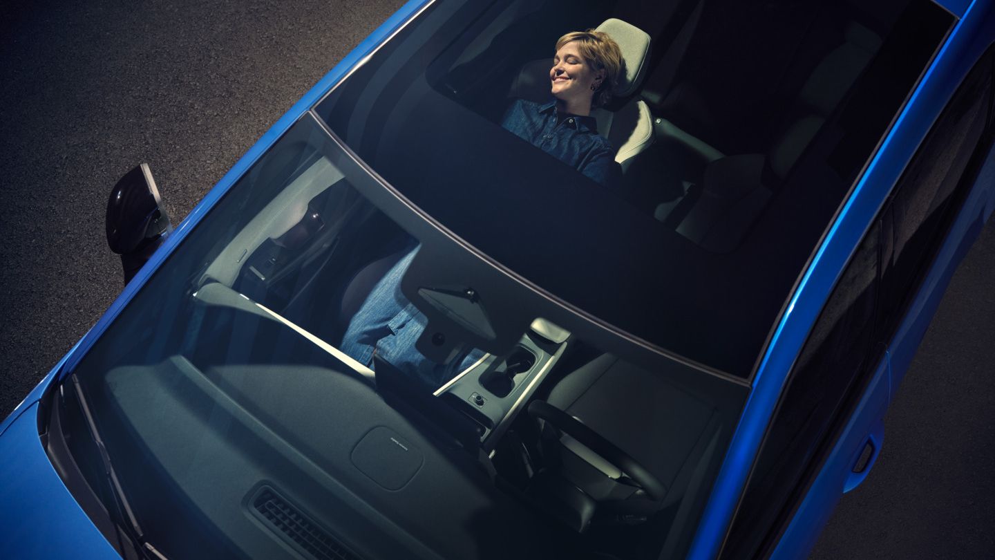 Blick von oben durch das Panoramadach eines blauen VW ID. Polo Style. Auf dem zurückgelehnten Beifahrersitz entspannt eine Frau.