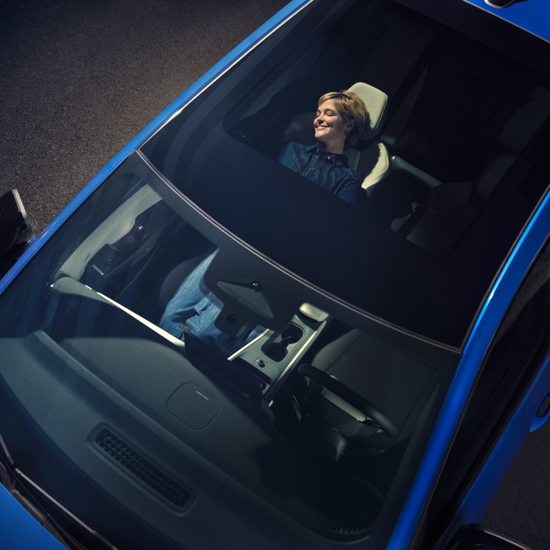 Blick von oben durch das Panoramadach eines blauen VW ID. Polo Style. Auf dem zurückgelehnten Beifahrersitz entspannt eine Frau.