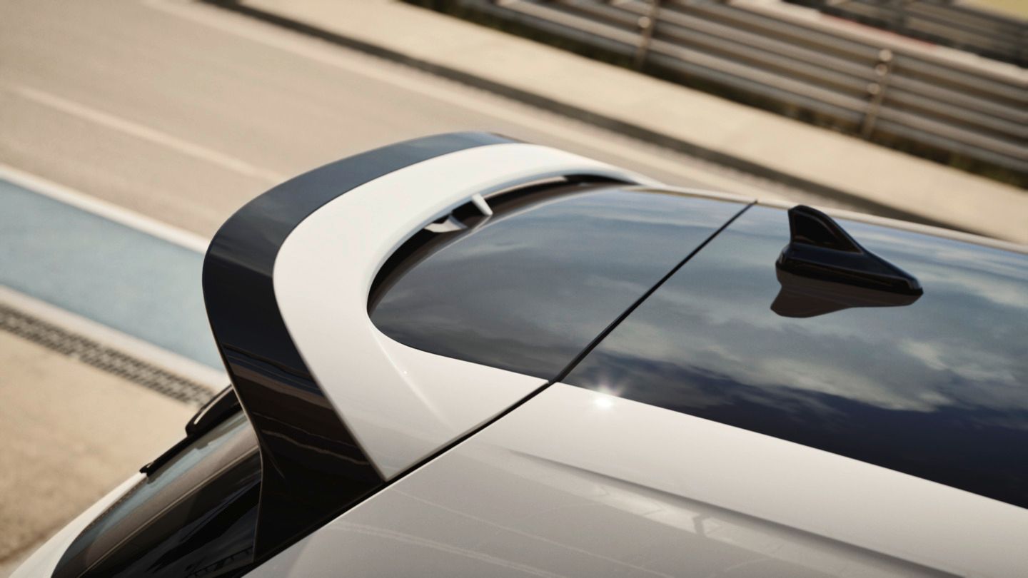 Nahaufnahme des Heckbereichs eines modernen Autos mit glänzendem schwarzem Dach und weißem Spoiler. Auf dem Dach ist eine kleine, haifischflossenartige Antenne zu sehen. Im Hintergrund erkennt man unscharf eine Rennstrecke oder Teststrecke mit Leitplanken.