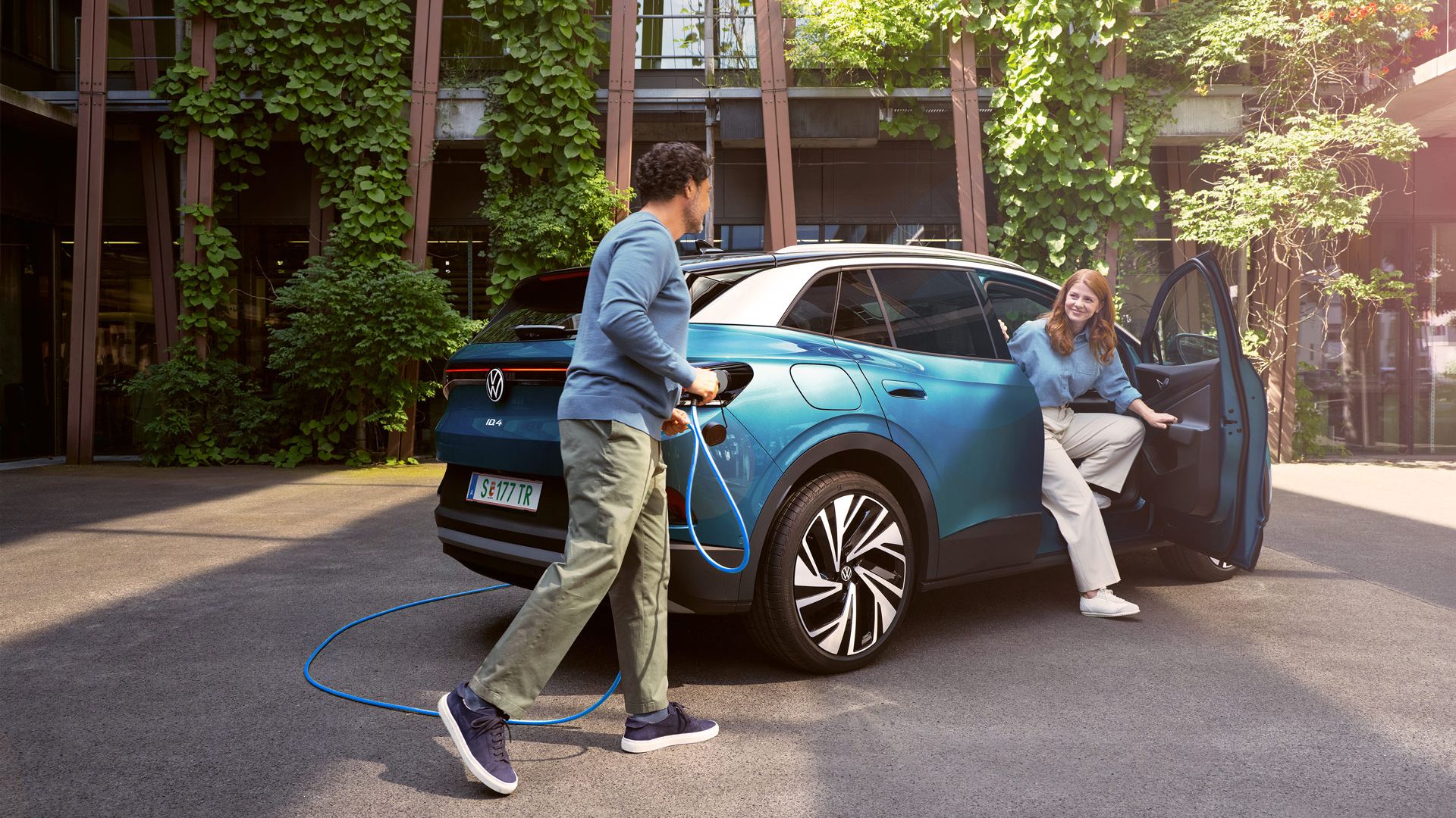 Eine Frau steigt aus einem blauem ID.4 aus, während ein Mann das POWER 2 Go Kabel anschließt. Im Hintergrund sind grüne Pflanzen und moderne Gebäude zu sehen.