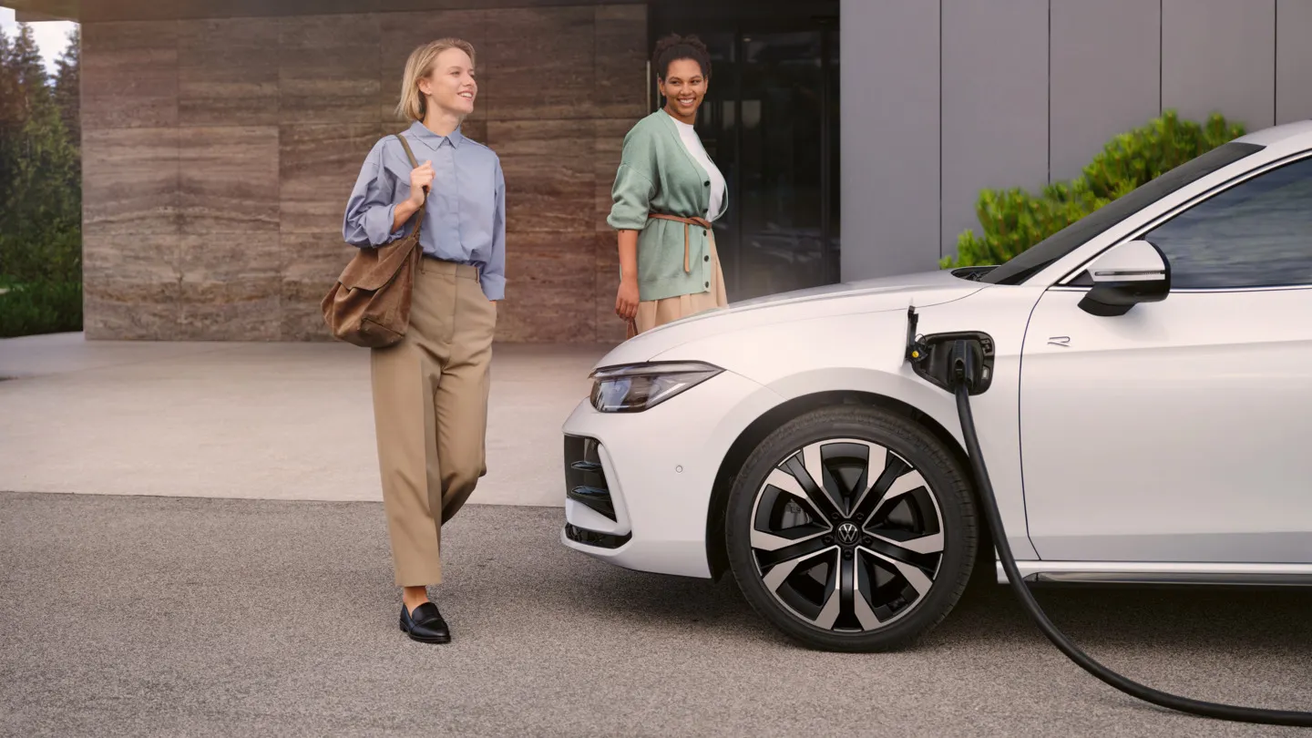 Zwei Frauen gehen auf einen weißen VW Passat R-Line eHybrid zu, der gerade geladen wird.