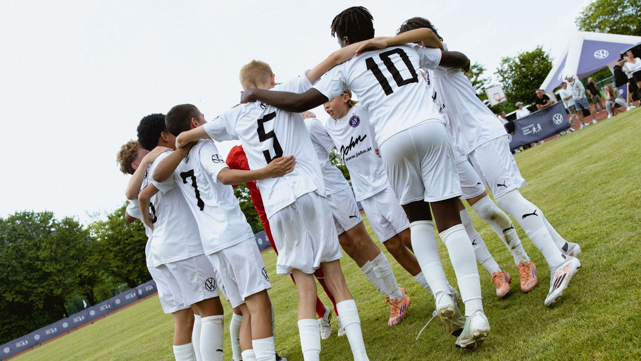 Eine Jugendfußballmannschaft in weißen Trikots steht im Kreis auf dem Spielfeld und legt die Arme umeinander. Die Spieler tragen Rückennummern, darunter gut sichtbar die Nummern 5 und 10. Im Hintergrund sind Bäume, ein Zaun mit Werbebannern und ein blauer Himmel mit ein paar Wolken zu sehen.