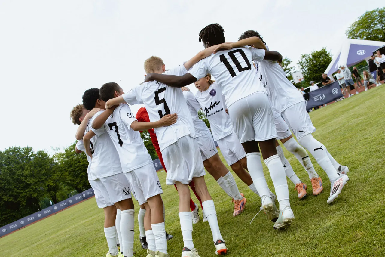 Eine Jugendfußballmannschaft in weißen Trikots steht im Kreis auf dem Spielfeld und legt die Arme umeinander. Die Spieler tragen Rückennummern, darunter gut sichtbar die Nummern 5 und 10. Im Hintergrund sind Bäume, ein Zaun mit Werbebannern und ein blauer Himmel mit ein paar Wolken zu sehen.
